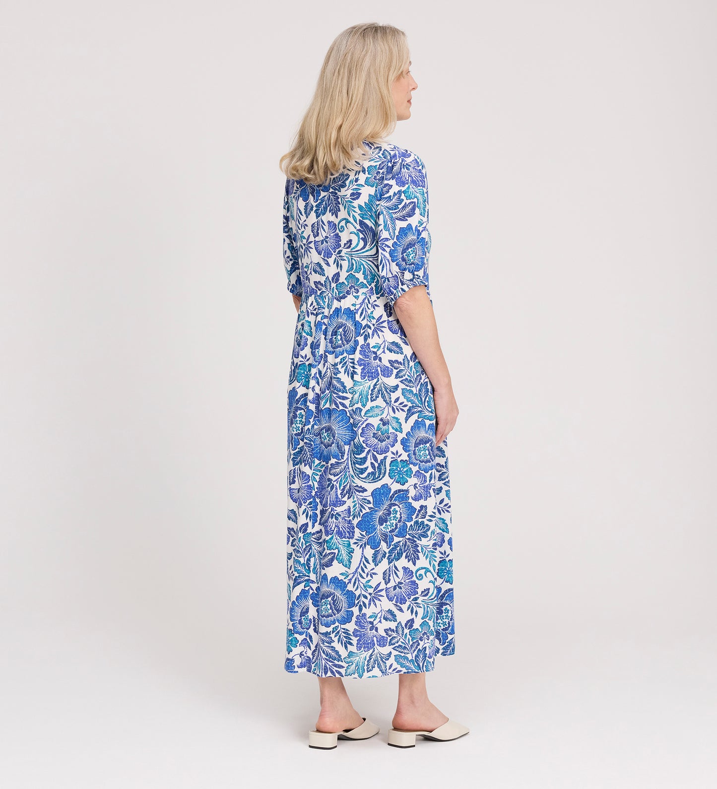 A woman with blonde hair stands sideways against a plain white background, wearing the Logan Blue Floral Midi Dress with a relaxed fit, blue and white floral print, elbow-length sleeves, and light-colored flat shoes.