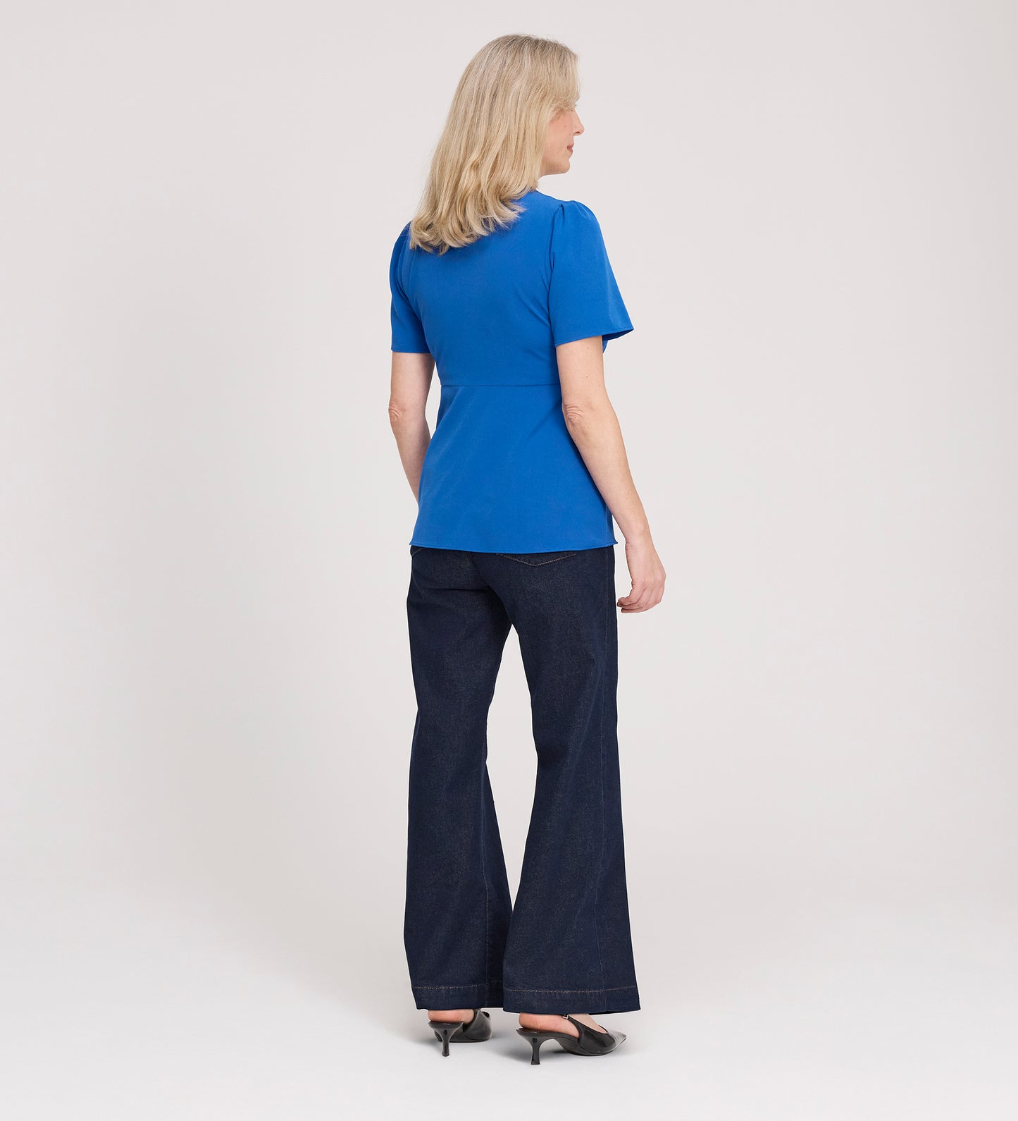 A woman with blonde hair faces away, wearing a Maryam Cobalt Button Up V-Neck Short Sleeve Top, dark flared jeans, and black heels, set against a plain white background.