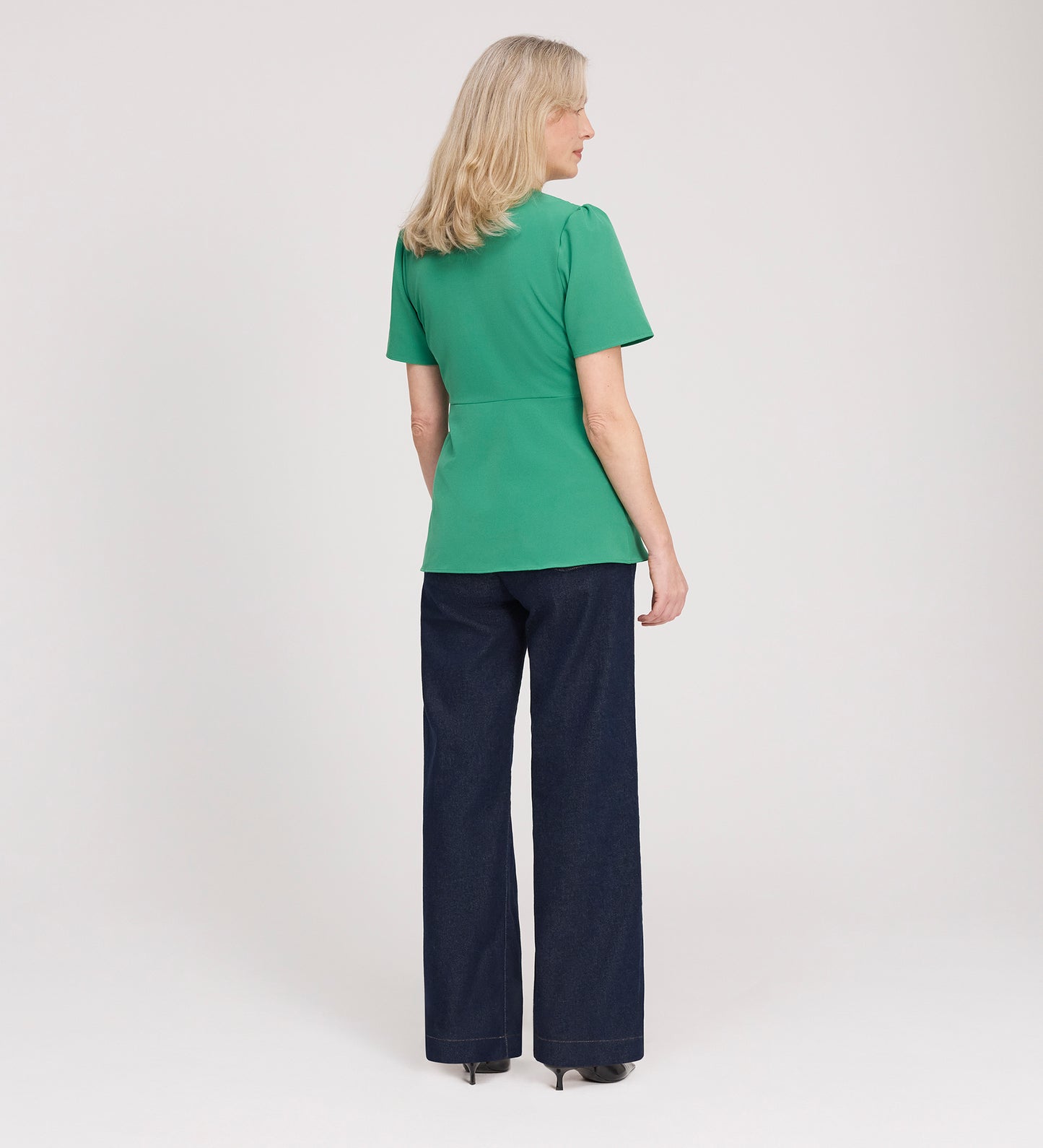 A woman with blonde hair faces away, wearing the Maryam Green Button Up V-Neck Short Sleeve Top, dark wide-leg pants, and heels against a plain white background.