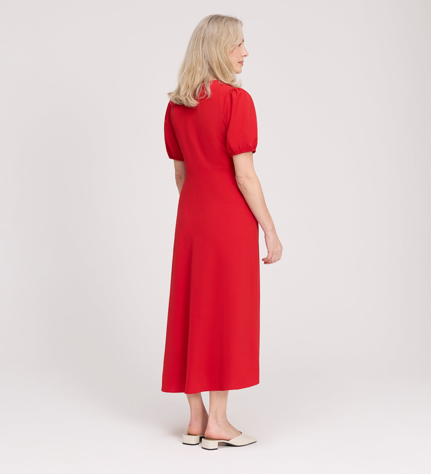 A woman with blonde hair stands facing away in a light studio, wearing the Robyn Red Button Through Midi Dress with short sleeves and beige slip-on shoes—an effortless look for day to evening.