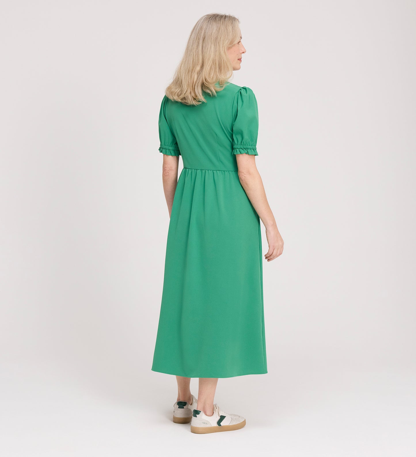 A woman with blonde hair stands facing away, wearing the Samantha Green Button Through Midi Dress and white sneakers with green accents against a plain light background.