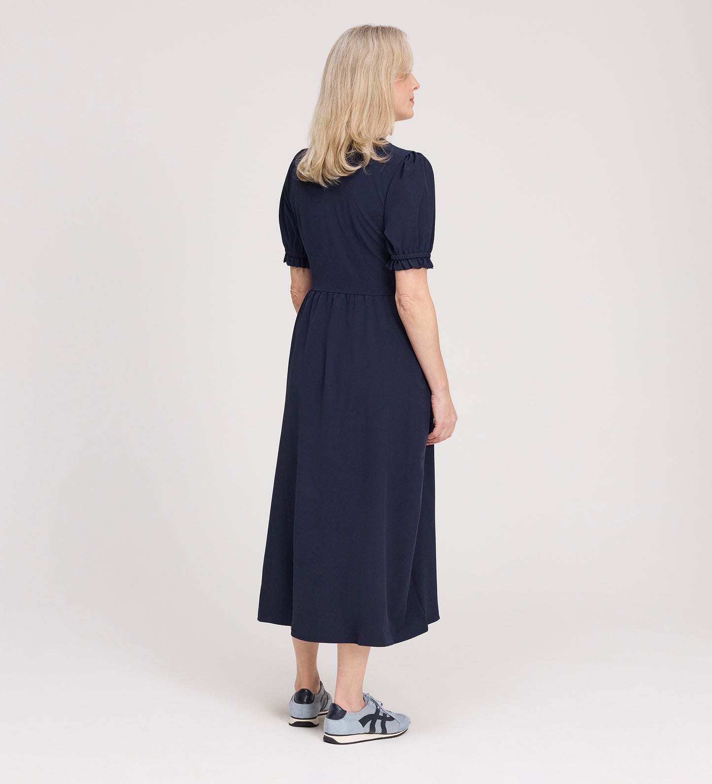 A woman with blonde hair faces away, wearing the Samantha Navy Button Through Midi Dress and gray sneakers with navy accents, set against a plain light background.