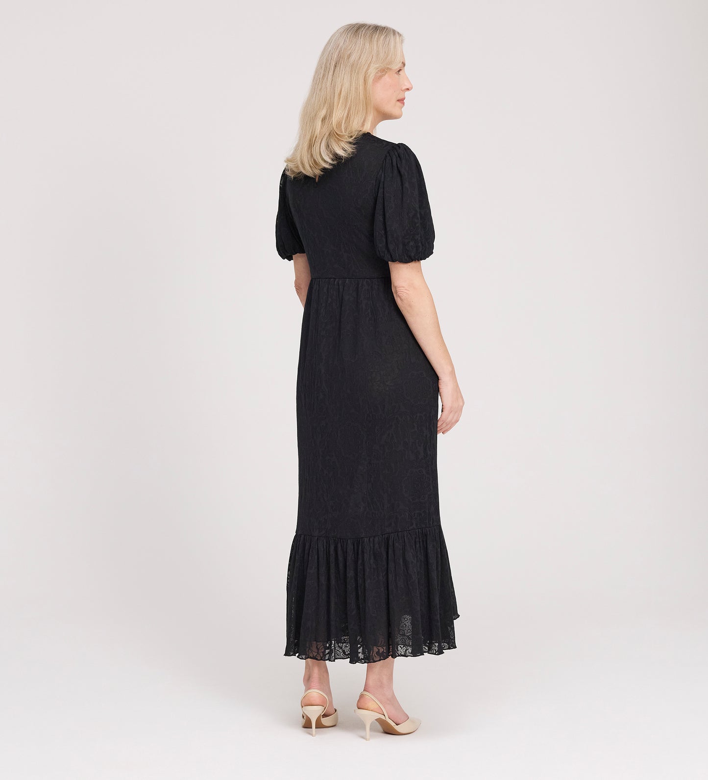 A woman with blonde hair faces away, wearing the Yasmine Black Jersey Midi Dress featuring puff sleeves and a ruffled hem, paired with beige heeled shoes against a plain light background.