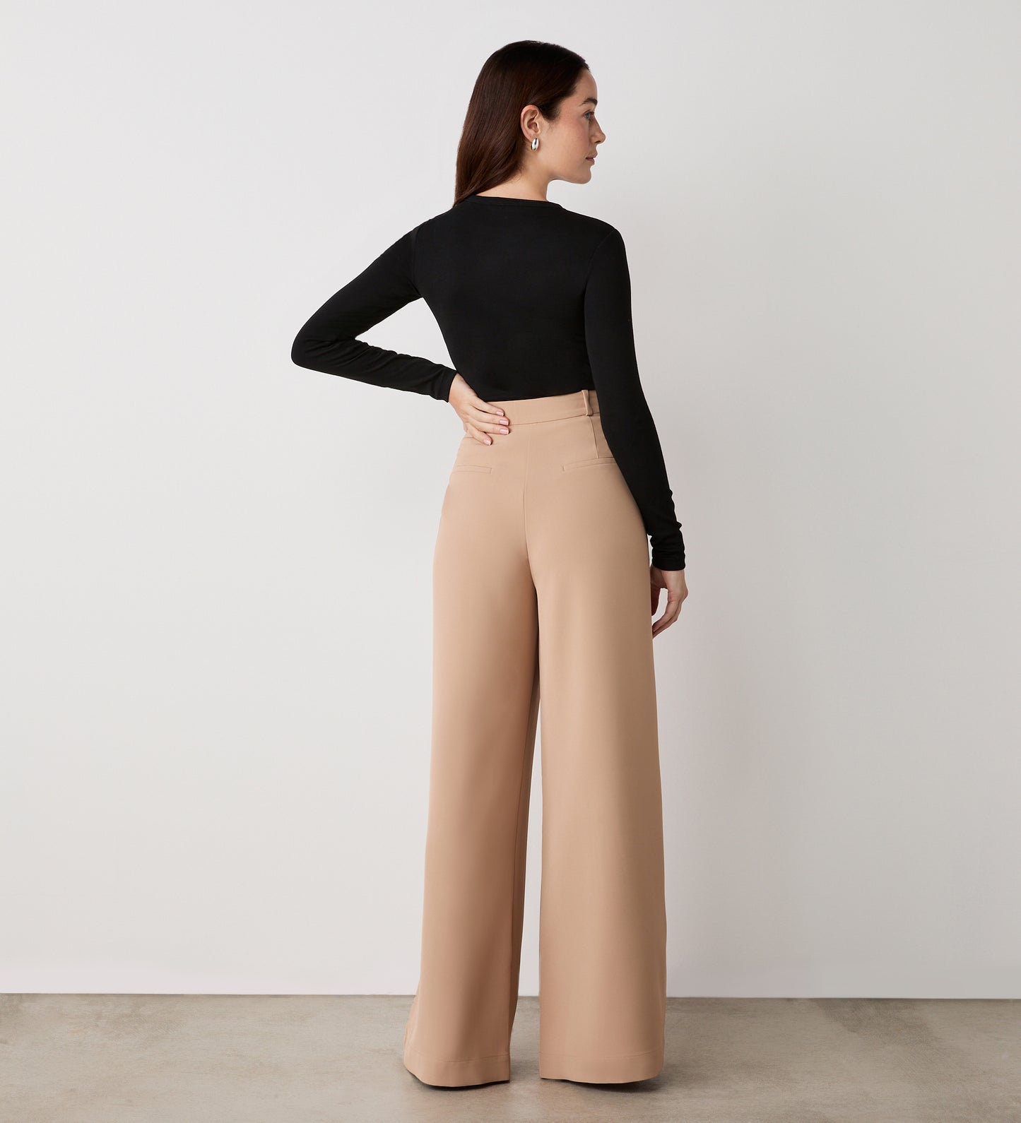 A woman stands facing away, wearing a fitted black long-sleeve top and the Addie Camel Wide Leg Trousers with side pockets, in a minimal indoor setting featuring a plain white wall and concrete floor.