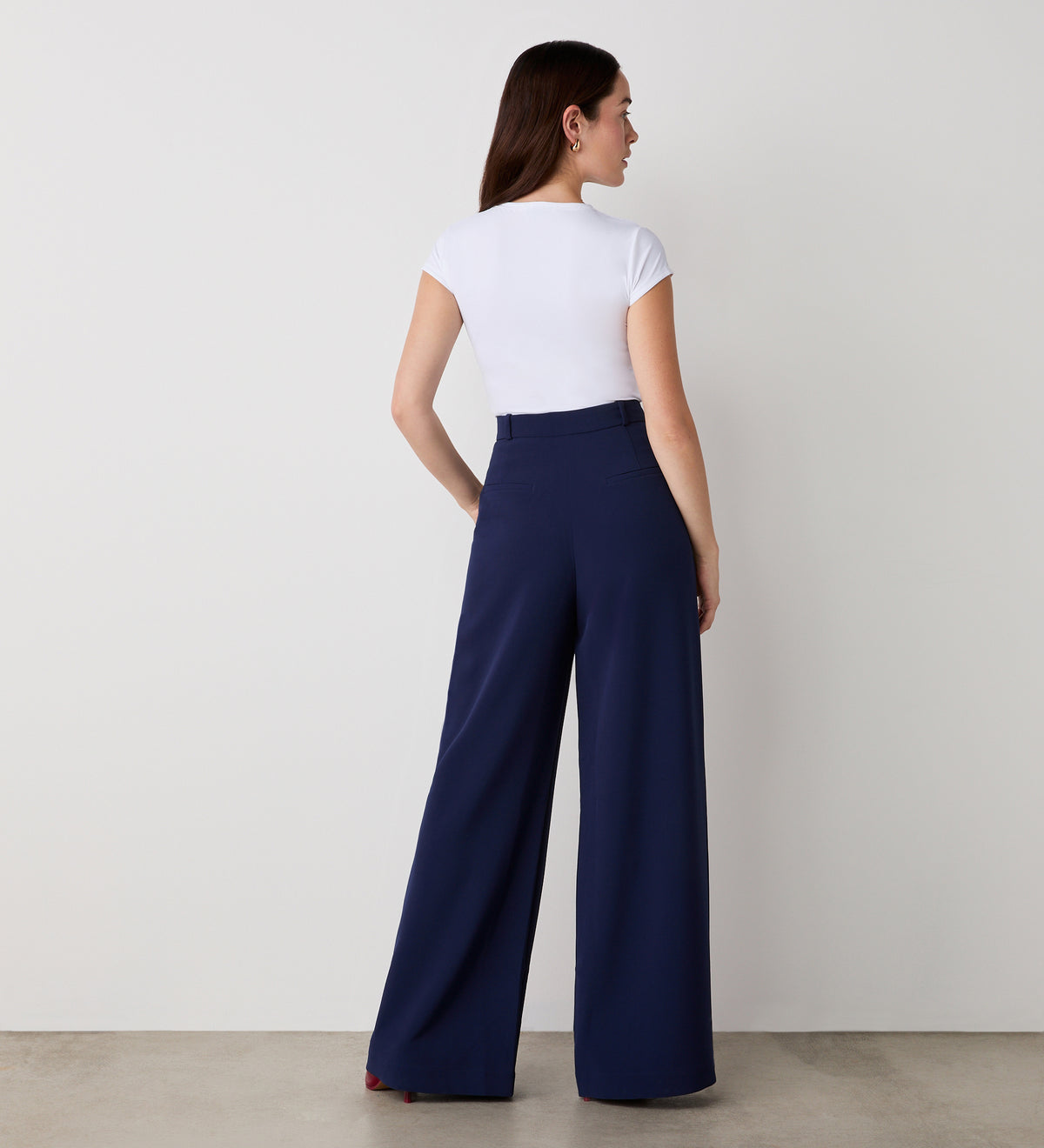 Woman stands with her back to the camera, wearing a fitted white short-sleeve top and Addie Navy Wide Leg Trousers, in front of a plain white wall and light-colored floor.