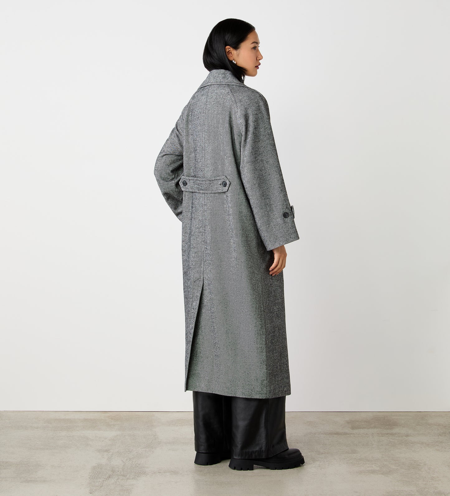 A woman with short dark hair models the Adrianna Grey Long Coat, a longline silhouette with a lapel collar, back belt detail, and center vent, layered over wide-leg black pants and chunky shoes against a white wall on concrete.