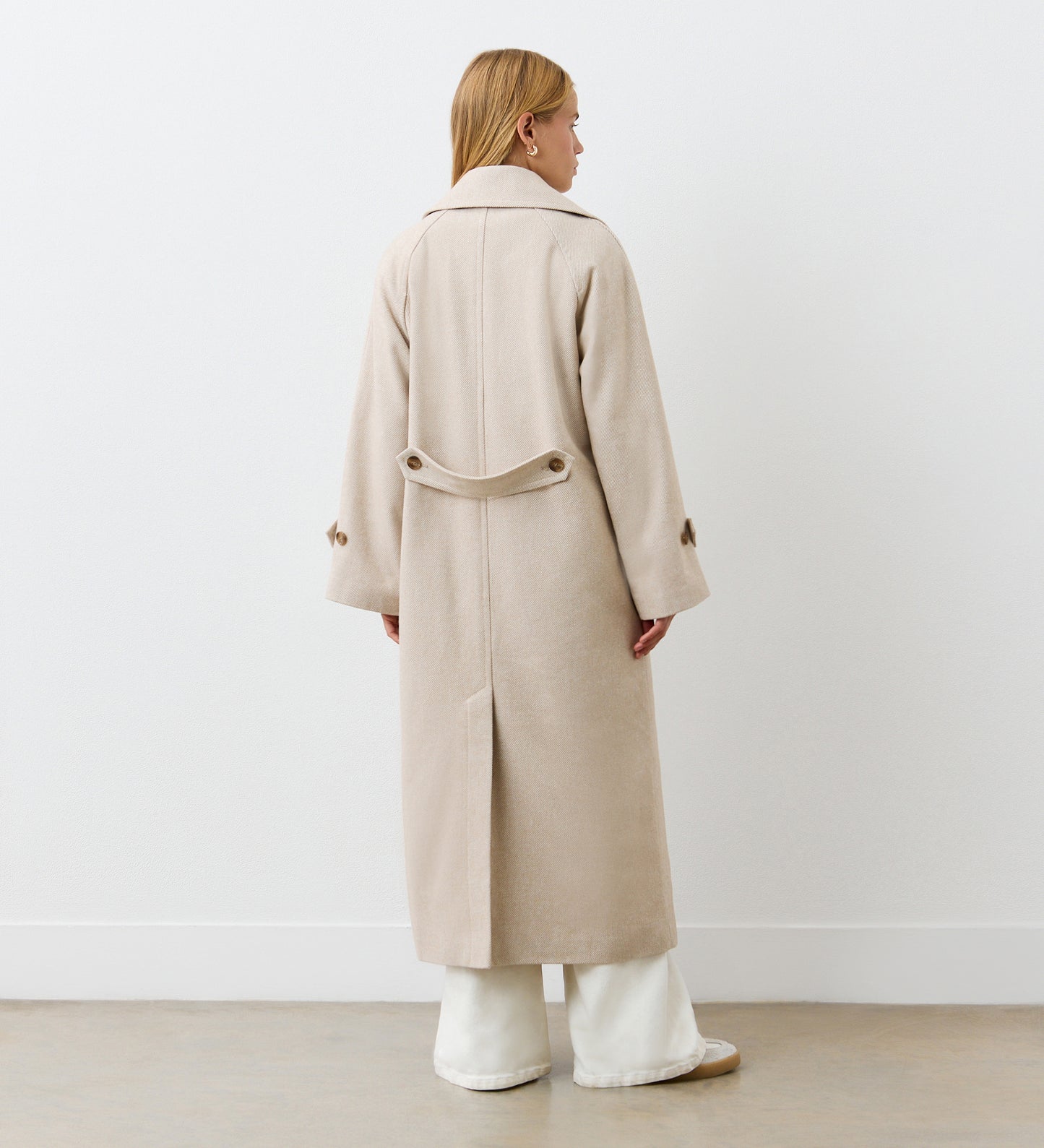 A person with blonde hair stands facing away, wearing the Adrianna Camel Long Coat with a classic lapel collar and back belt detail, layered over wide white pants, against a plain white wall and light-colored floor.