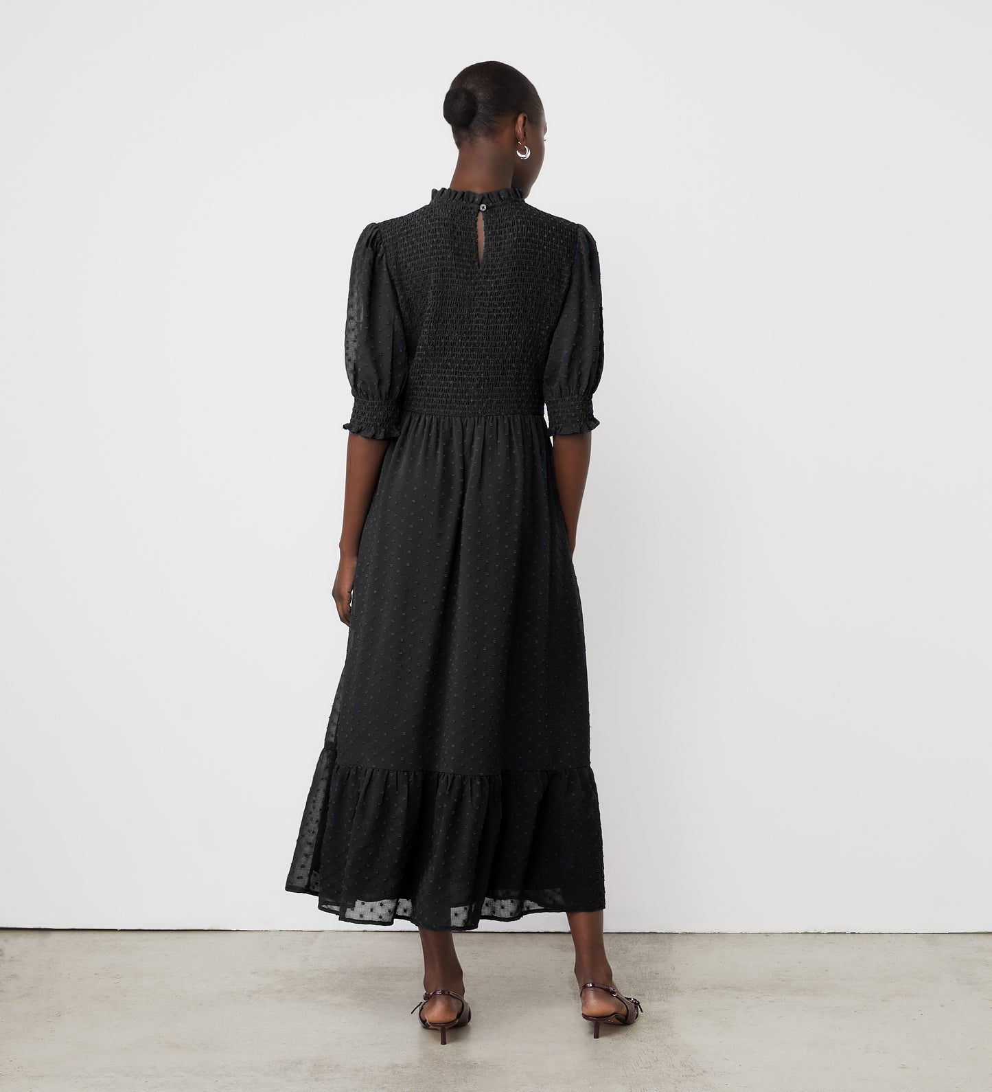 A woman with short hair faces a white wall, wearing the Amelia Black Georgette Midi Dress. The dress has puffed half sleeves, a high neckline, a shirred bodice, ruffled hem, and she pairs it with brown sandals.