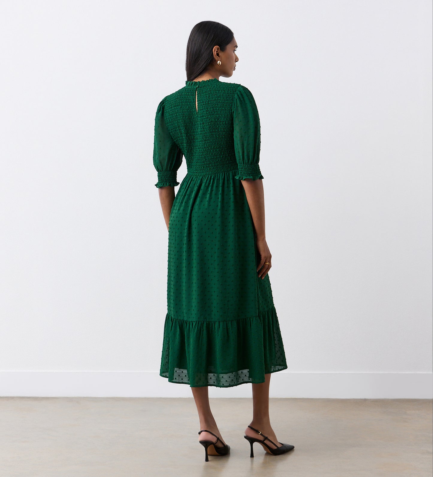 A woman with medium-length dark hair wears the Amelia Dark Green Georgette Midi Dress, featuring puffed half sleeves, a shirred bodice, and a tiered hemline. She styles it with black ankle-strap heels and stands facing away.