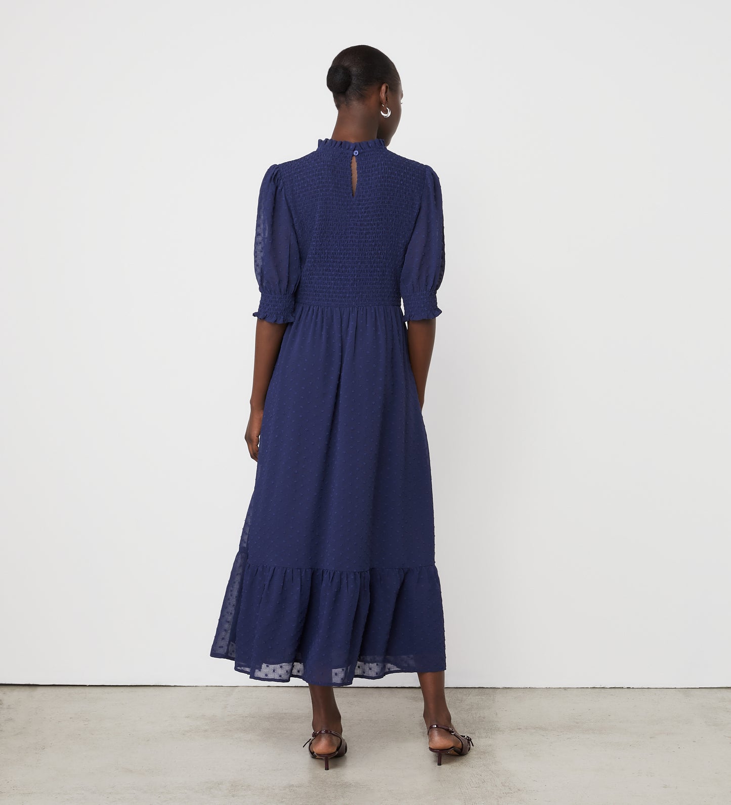 A woman with short hair and sandals stands on a light concrete floor against a white wall, wearing the Amelia Navy Chiffon Midi Dress featuring puff sleeves, textured chiffon fabric, and a tiered midi silhouette.