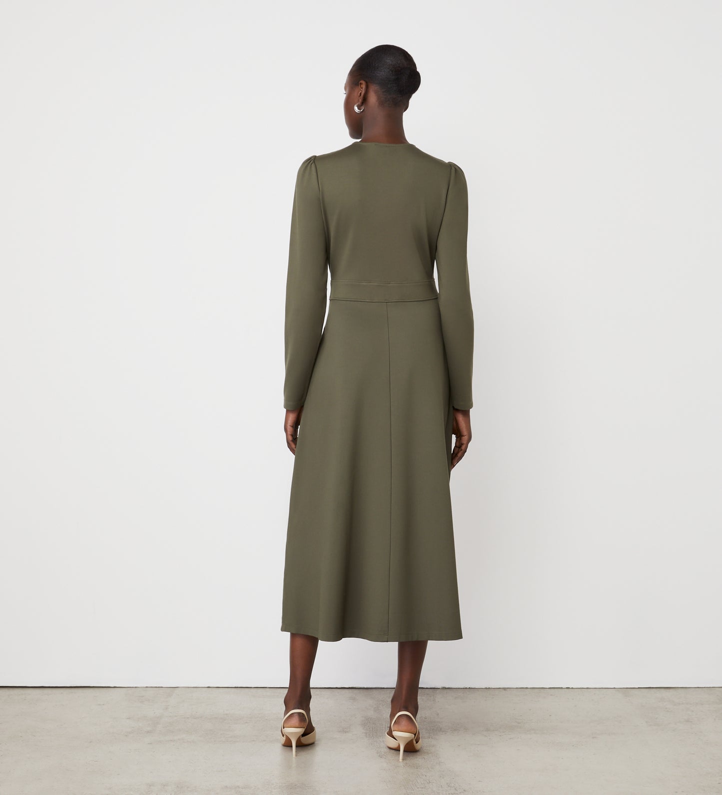 A woman faces away from the camera, wearing the Aria Khaki Ponte Jersey Midi Dress with an empire waistband and light heels, standing against a plain white wall and concrete floor.