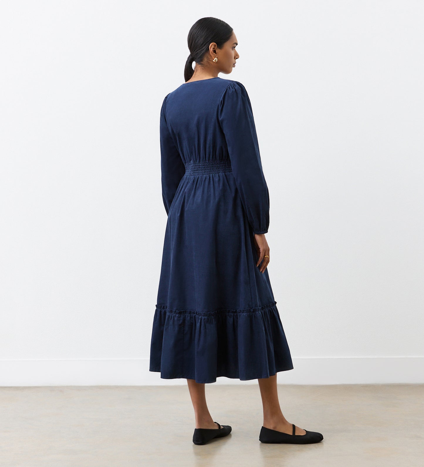 A woman with her hair tied back stands in a minimal, light-walled room, facing away and wearing the Arizona Navy Corduroy Midi Dress with a flared hem, paired with black flats.