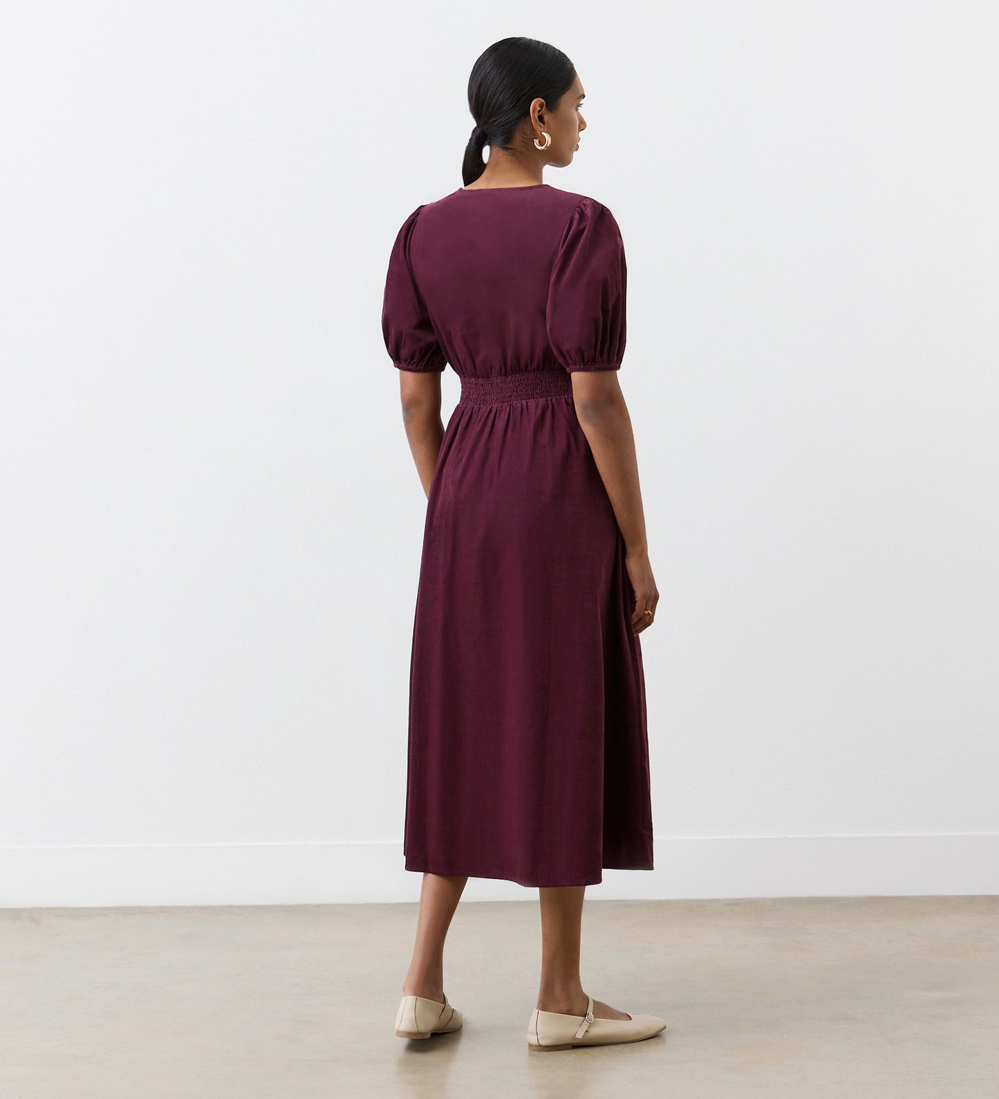 A woman with short hair tied back wears the Arlow Burgundy Corduroy Midi Dress, featuring short puff sleeves and an elastic waist—perfect for autumn—paired with beige flats, standing indoors against a plain white wall and floor.