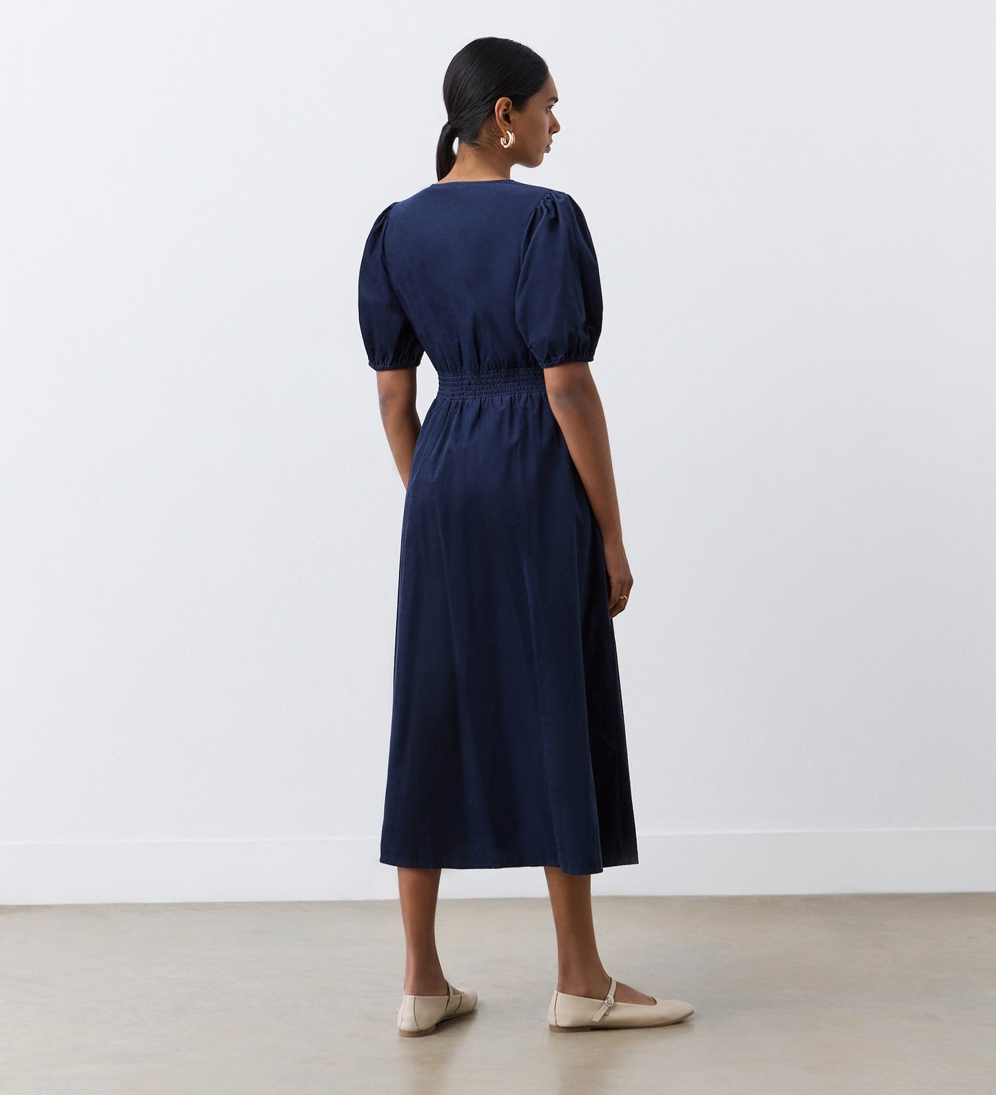 A woman with dark hair in a low ponytail, wearing the Arlow Navy Corduroy Midi Dress with elasticated puff sleeves and beige flat shoes, stands facing away against a plain white wall and light brown floor.