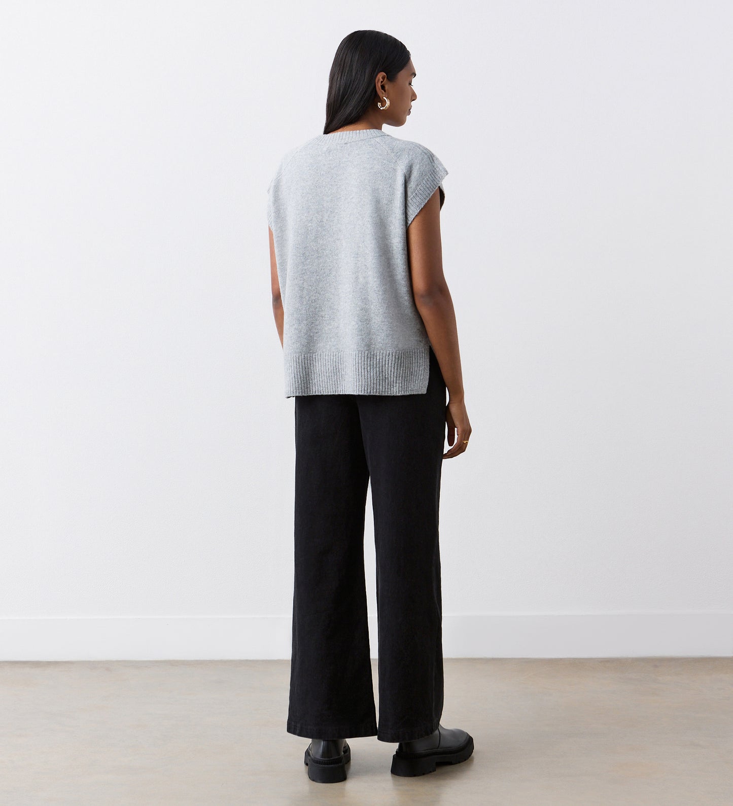 A person with long dark hair faces away, wearing an Ava Grey Knitted Vest over a short-sleeved top, paired with black wide-leg pants and black shoes against a white wall—an ideal autumn layering look.