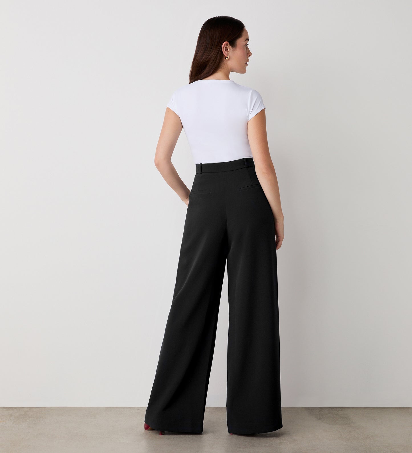 A woman stands facing away from the camera, wearing a fitted white short-sleeve top tucked into the Addie Black Wide Leg Trousers with functional side pockets, her straight brown hair set against a neutral background.