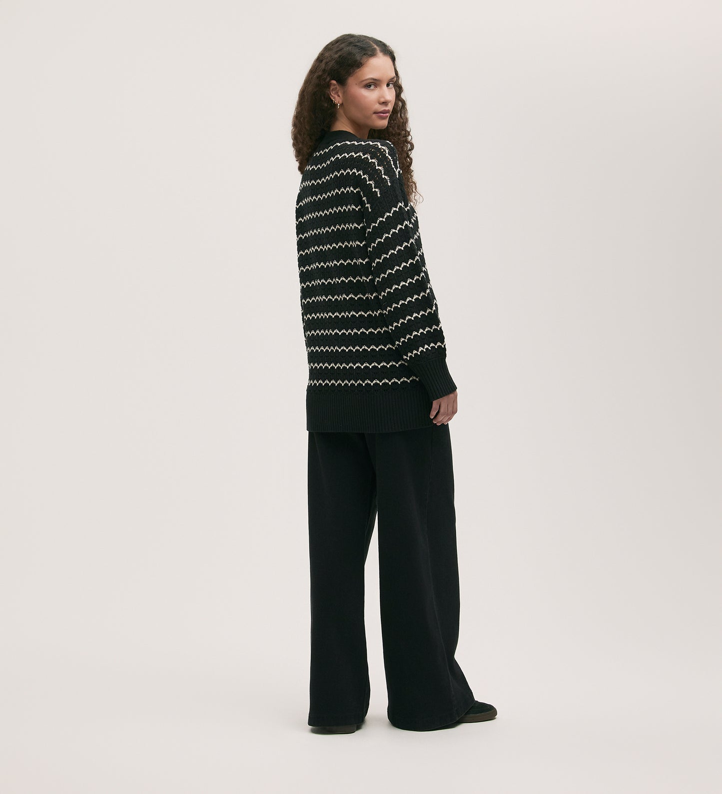 A person with long curly hair stands sideways, looking back at the camera, wearing the Alena Black Crochet Cardigan and loose black pants against a plain light background.