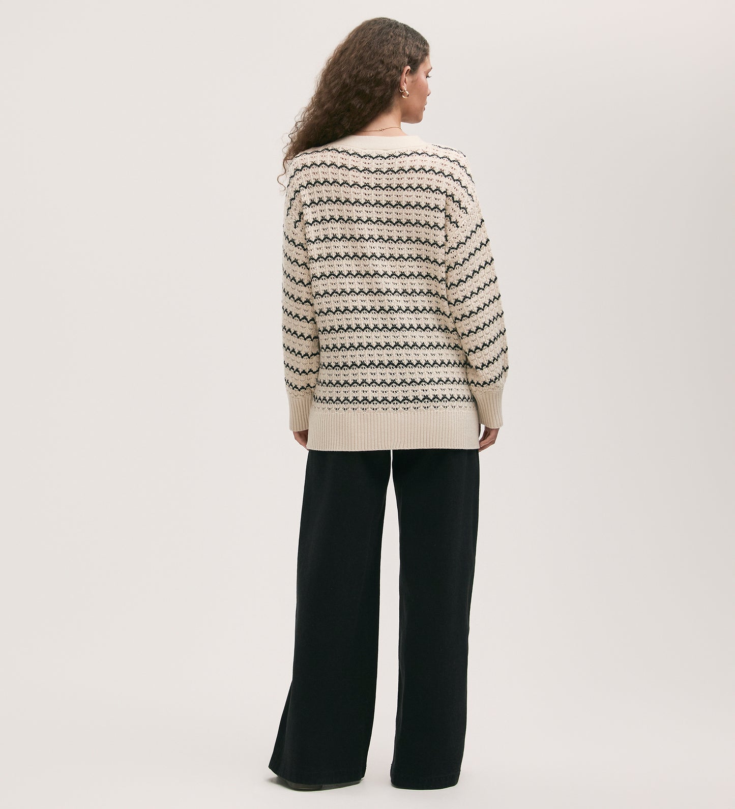 A woman with long curly hair, seen from the back, wears the oversized Alena Ivory Crochet Cardigan and loose black pants against a plain light background.