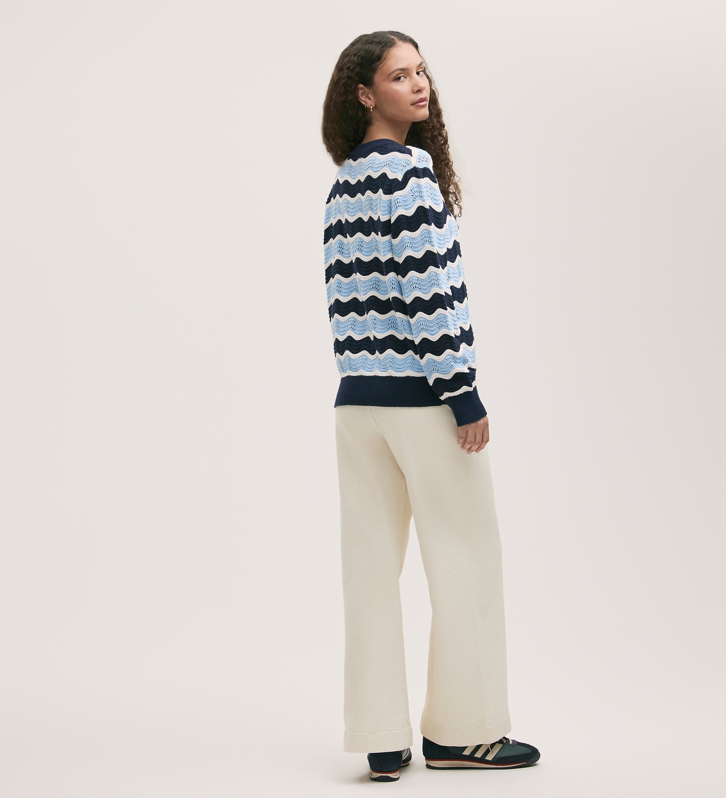 A person with long curly hair wears the Antonia Blue Stripe Crochet Jumper, cream wide-leg pants, and black sneakers with beige stripes, standing and looking over their shoulder against a plain light background.