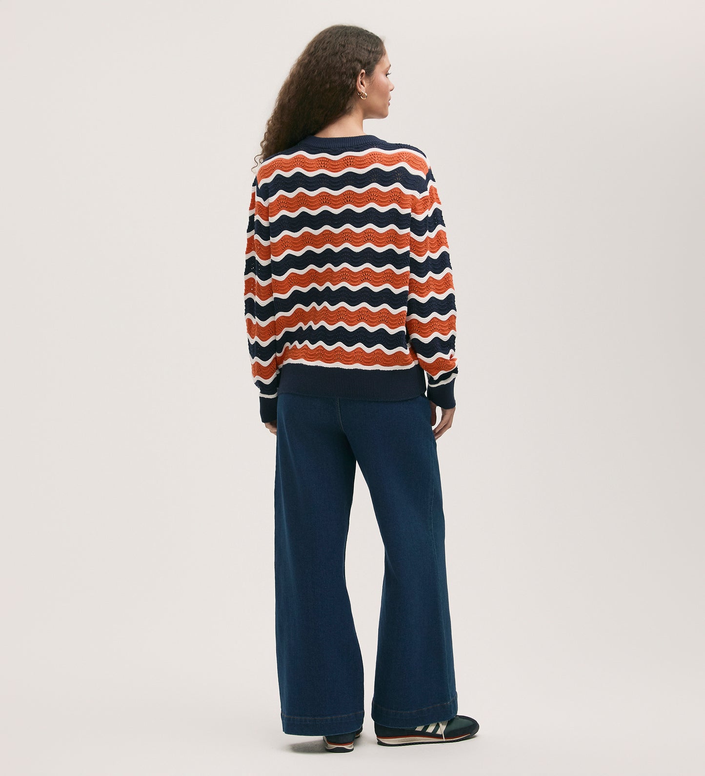 A person with long curly hair faces away, wearing the Antonia Orange Stripe Crochet Jumper in a relaxed fit with wavy stripes of red, orange, white, and navy, paired with wide-leg blue jeans and dark shoes against a plain background.