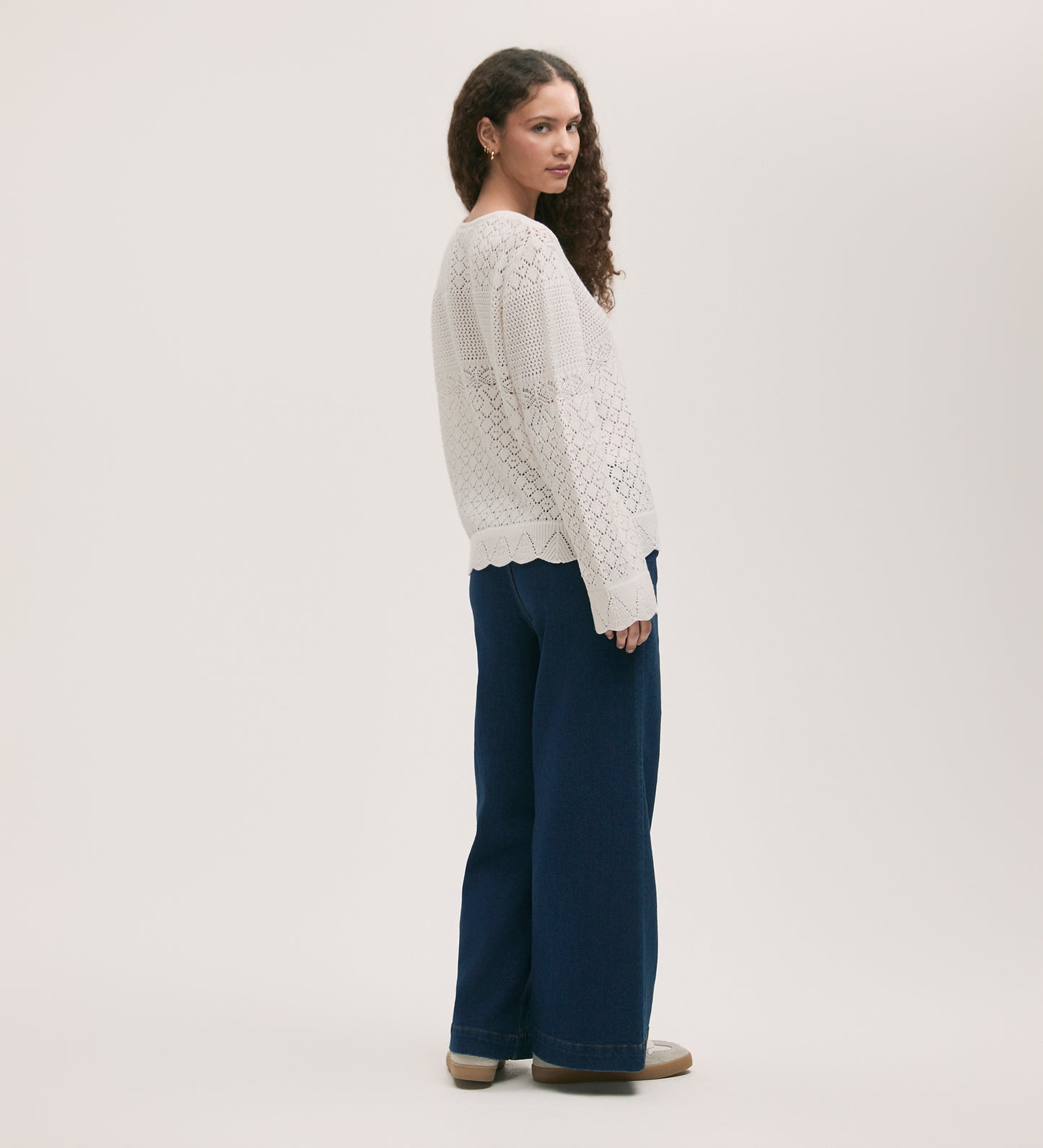 A woman with curly hair stands sideways against a light background, wearing the Audrenia Ivory Crochet Top, featuring scallop ribbed detailing, paired with wide-leg dark blue jeans and beige shoes.
