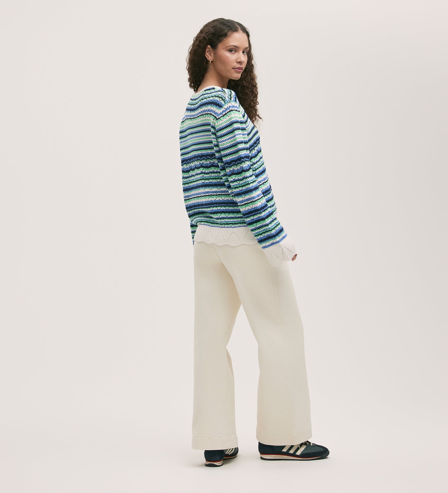 A woman with curly hair stands sideways against a plain background, wearing the Audrenia Blue Stripe Crochet Top, white wide-leg pants, and black sneakers with white stripes.