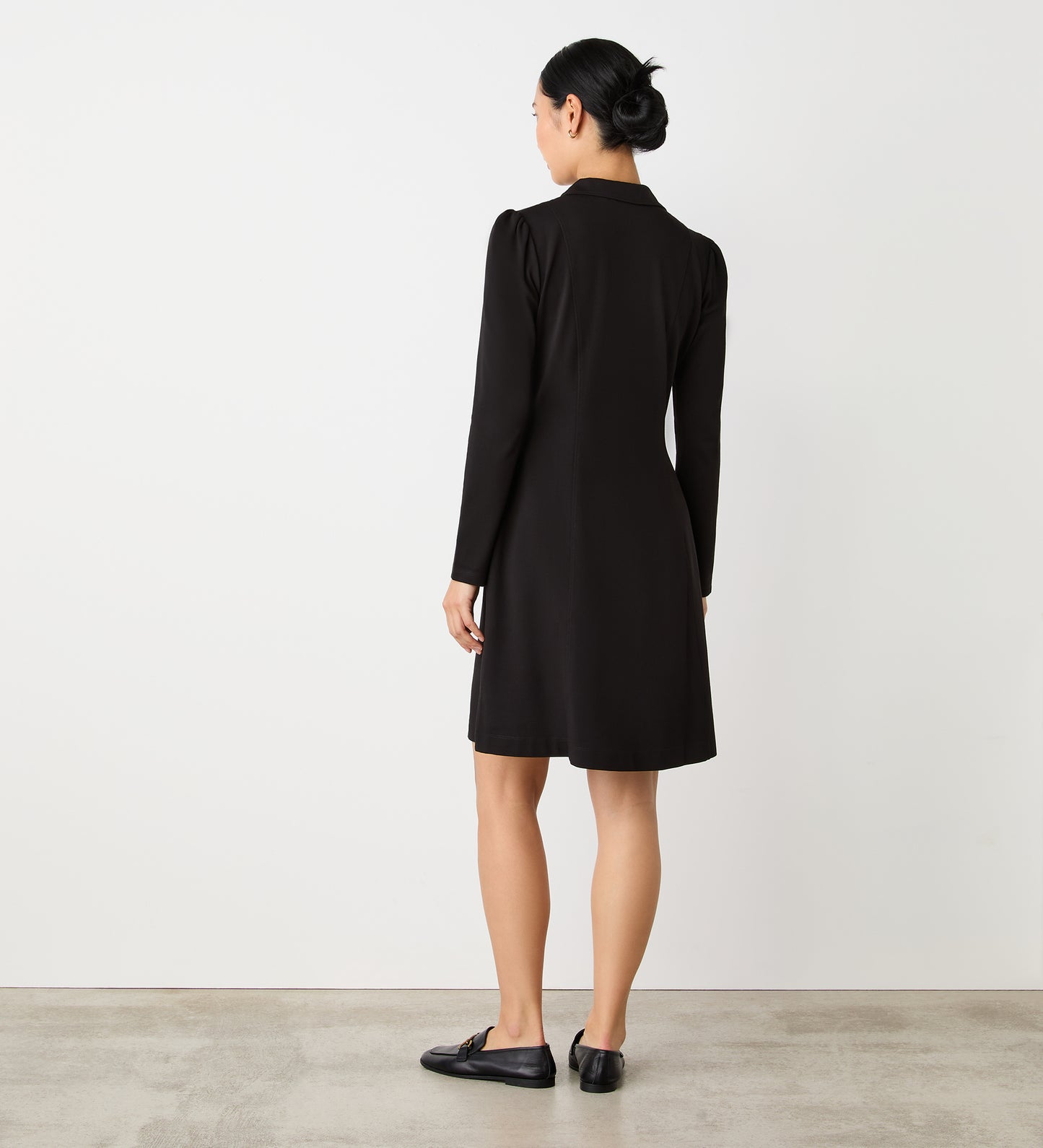 A woman with her hair in a low bun stands indoors, facing away from the camera, wearing the Bethany Black Ponte Jersey Midi Dress and black flats. The background is plain white with a concrete floor.