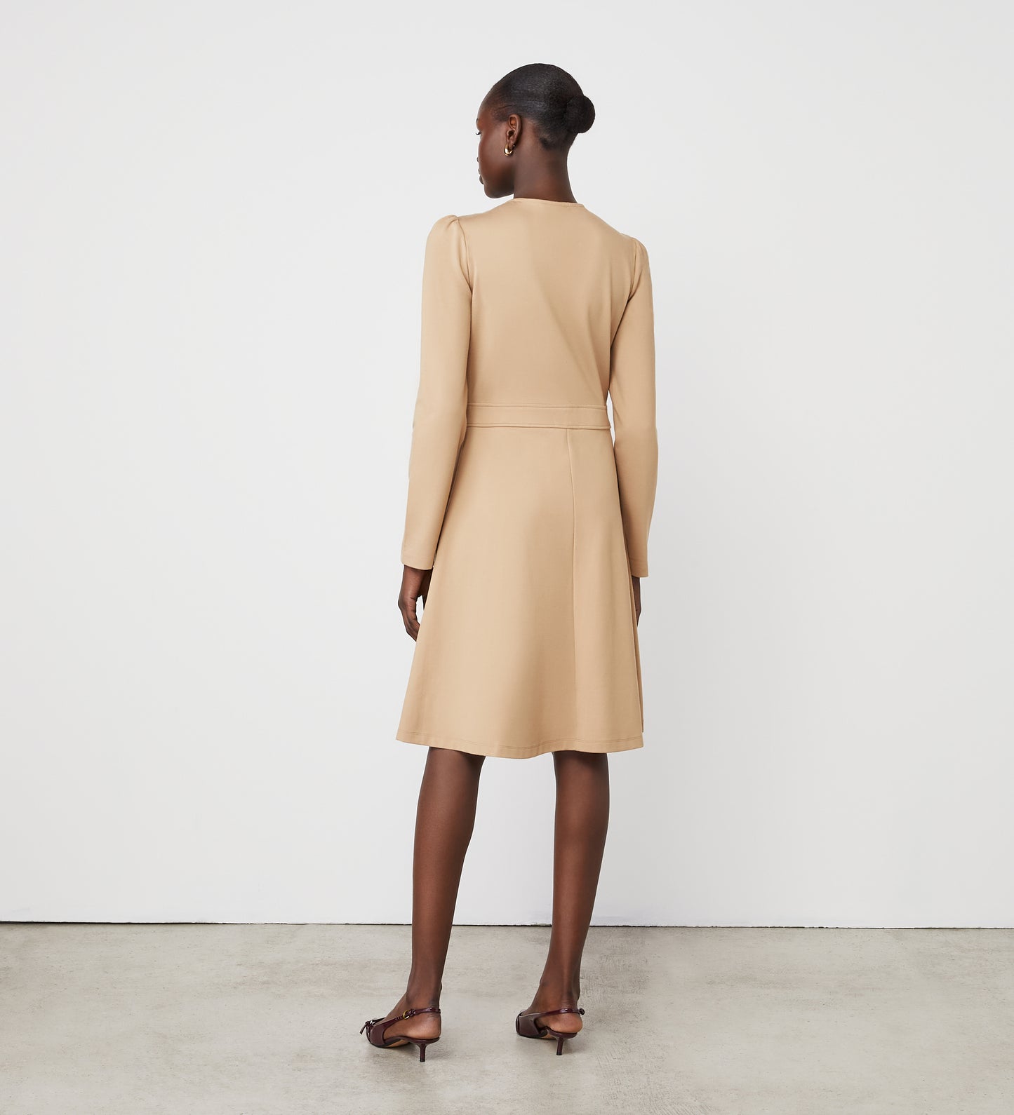 A woman faces away, wearing the Blake Camel Ponte Jersey Midi Dress and brown heeled sandals. Her hair is styled in a neat bun. The background is plain white with a gray concrete floor.