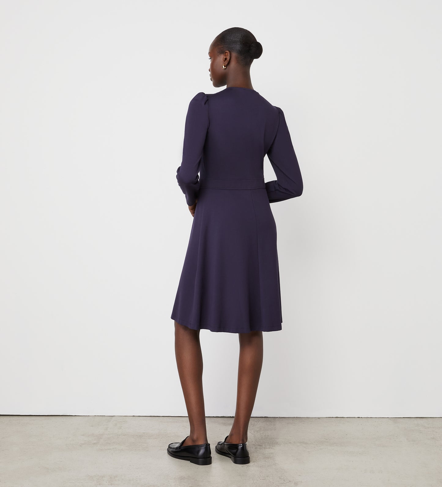 A woman faces away from the camera in the Blake Navy Ponte Jersey Midi Dress and black loafers. Her hair is tied back as she stands against a plain white wall on a concrete floor.