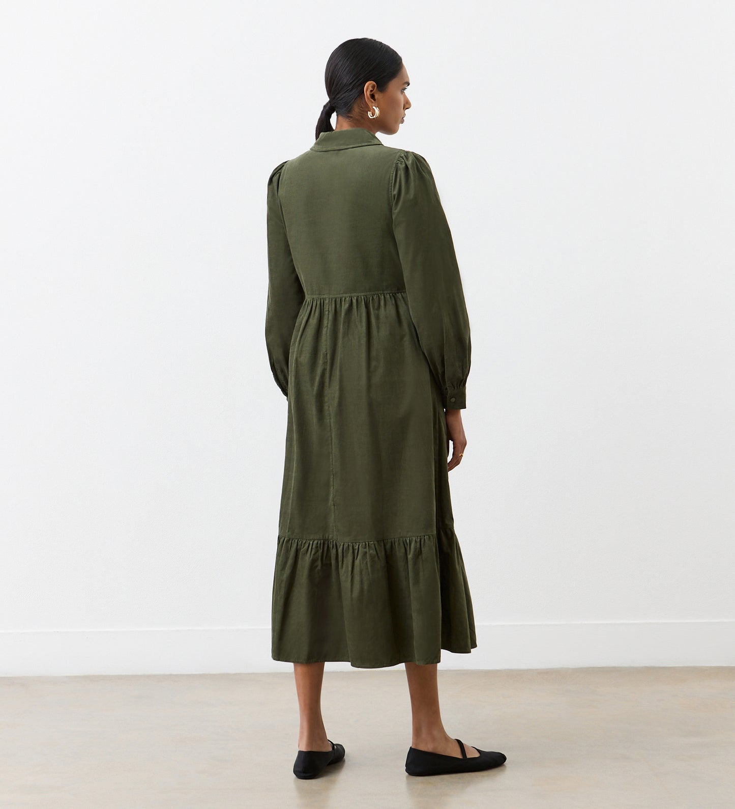 A woman with dark hair tied back, wearing the Callie Khaki Corduroy Midi Dress with long sleeves and a tiered hemline, stands indoors in front of a white wall facing away. She pairs the dress with black flat shoes.