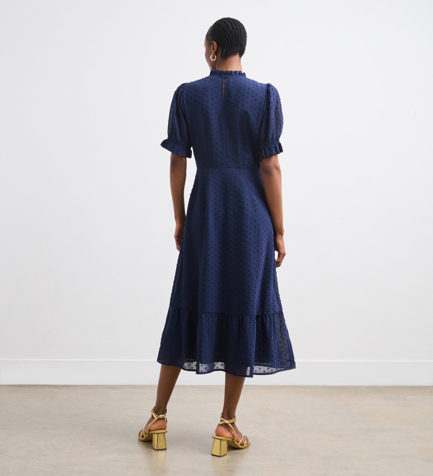 A woman with short hair and gold hoop earrings faces away, wearing the Camille Navy Chiffon Midi Dress with puff sleeves, ruffle neck, and tiered hem. She pairs it with tan heeled sandals and stands against a plain white background.