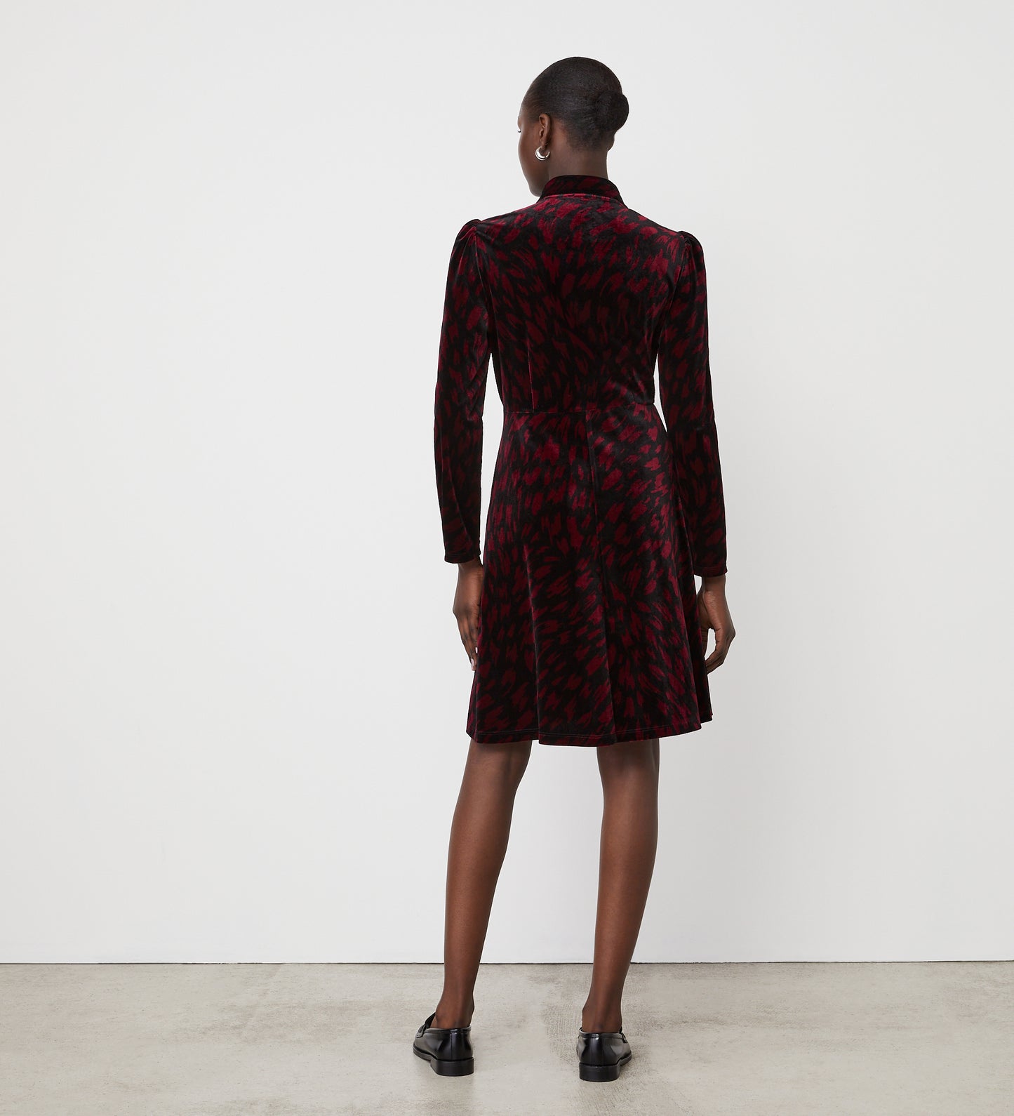 A person faces away from the camera wearing the Capri Burgundy Animal Knee Length Dress and black shoes, standing against a white wall and concrete floor—an ideal look for party season.