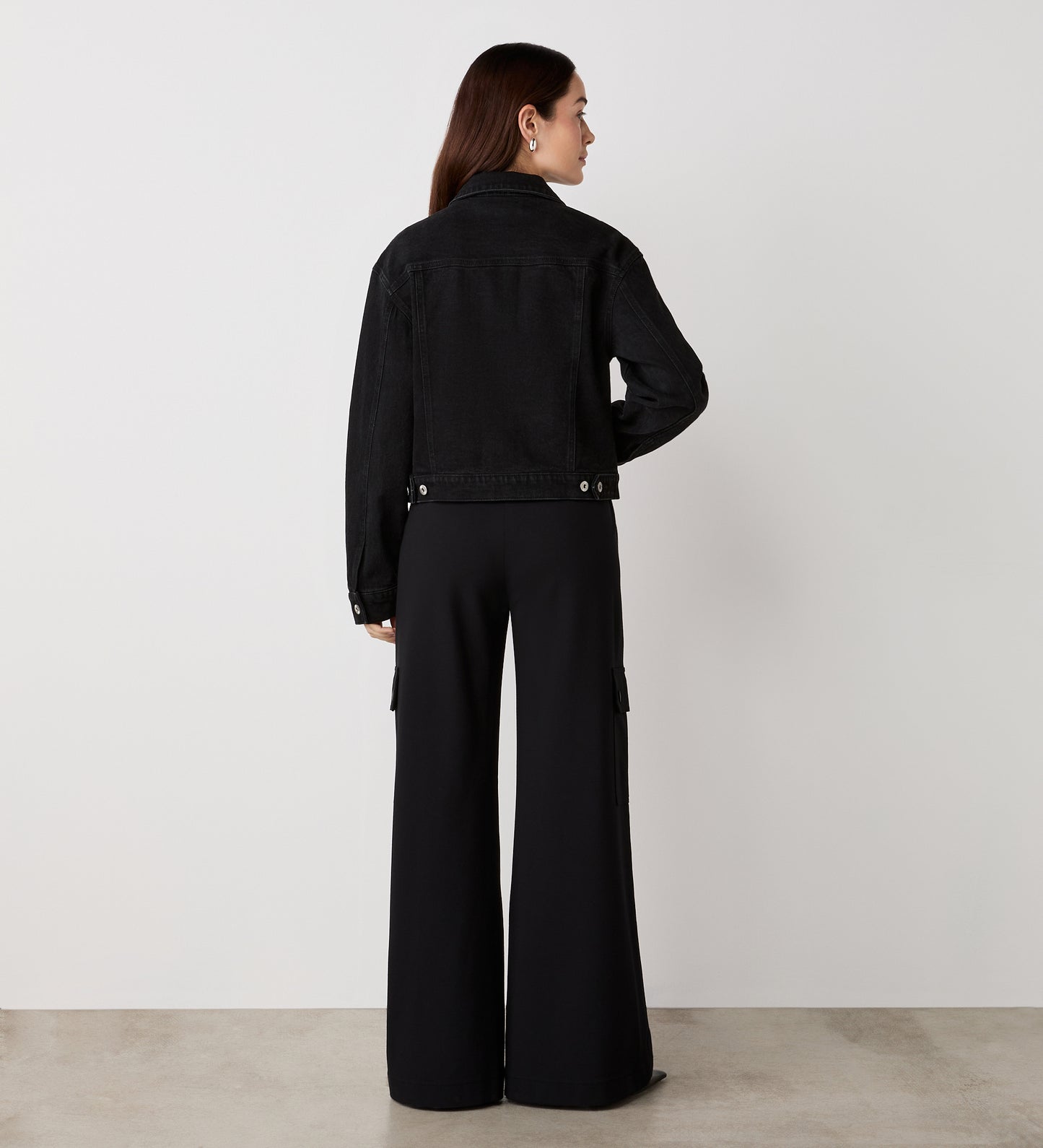 A woman with long brown hair stands in a minimal, light room, wearing the Caroline Black Denim Jacket with front patch pockets and black wide-leg pants, facing away from the camera on a smooth floor.