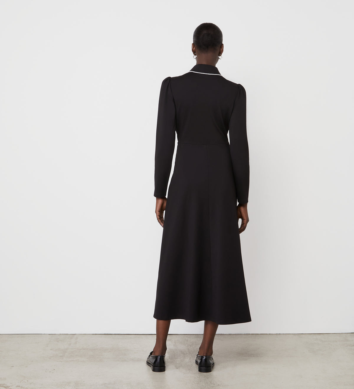 A person stands with their back to a white wall, wearing the Clarice Black Ponte Jersey Midi Dress featuring long sleeves, a fitted waist, slight puff shoulders, and a functional placket for added detail.