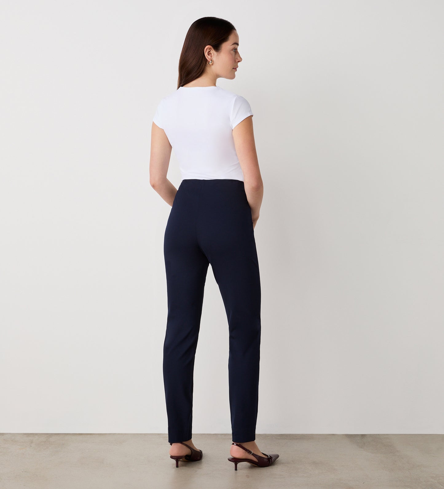 A woman with straight brown hair, wearing a fitted white short-sleeve top, Cleo Navy Ponte Jersey Leggings, and brown heeled shoes, stands facing a plain white wall on a light-colored floor.