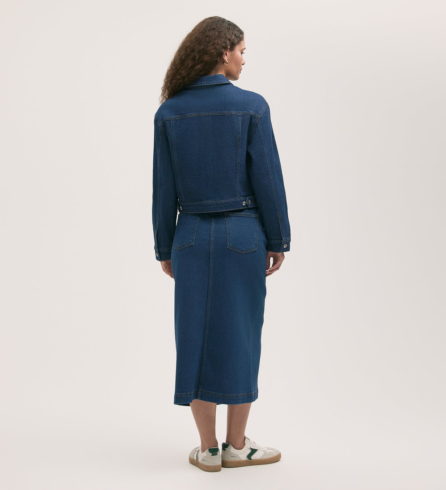 Wearing the Caroline Mid Wash Denim Jacket with front patch pockets and a matching midi skirt, a person with long, curly hair faces away in white sneakers against a plain light background—a true wardrobe essential.