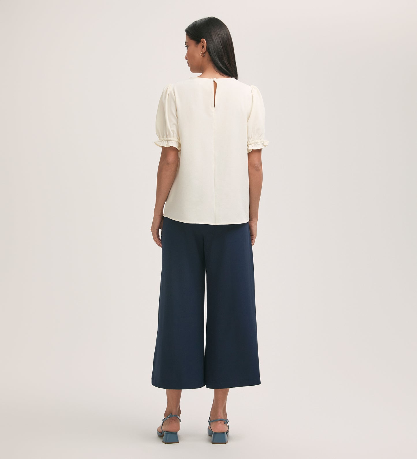 A woman faces away, wearing the Charlie Ivory Crepe T-Shirt with puff sleeves and back keyhole, navy wide-leg pants, and blue heeled sandals against a plain light background.