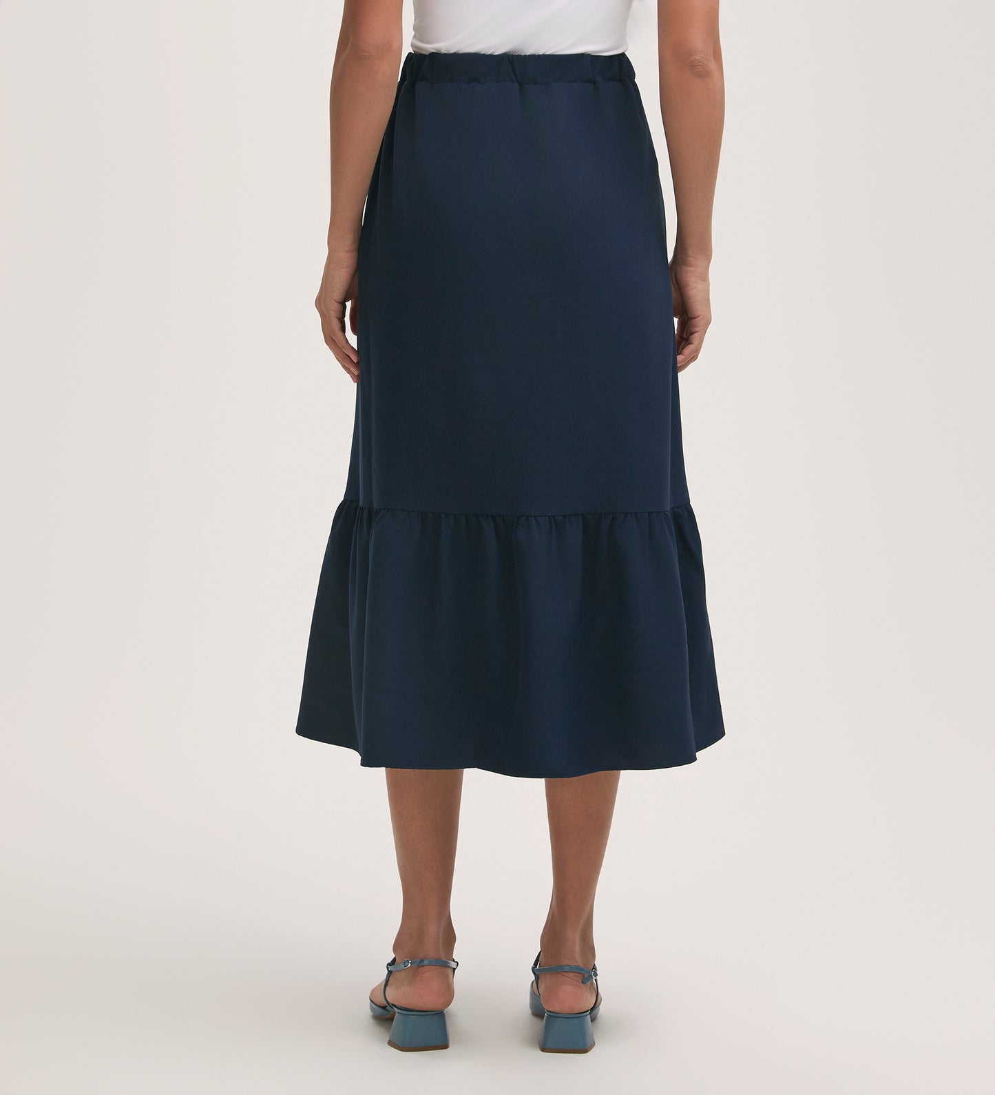 A person shown from the back against a plain light background wears a white top and the Riley Navy Crepe Tiered Midi Skirt with ruffled hem, elasticated waistband, and crease-free fabric, paired with blue heeled sandals.