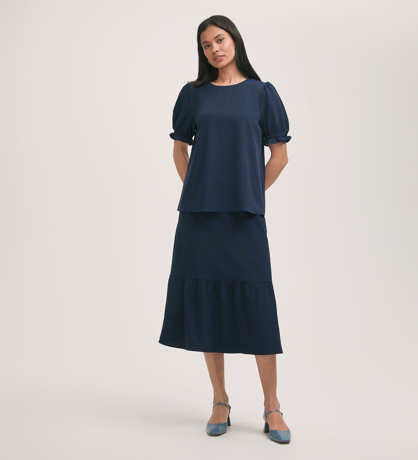 A woman stands against a plain light background in the Charlie Navy Crepe T-Shirt with puff sleeves and a matching midi skirt. She has straight dark hair and finishes her look with blue low-heeled shoes.