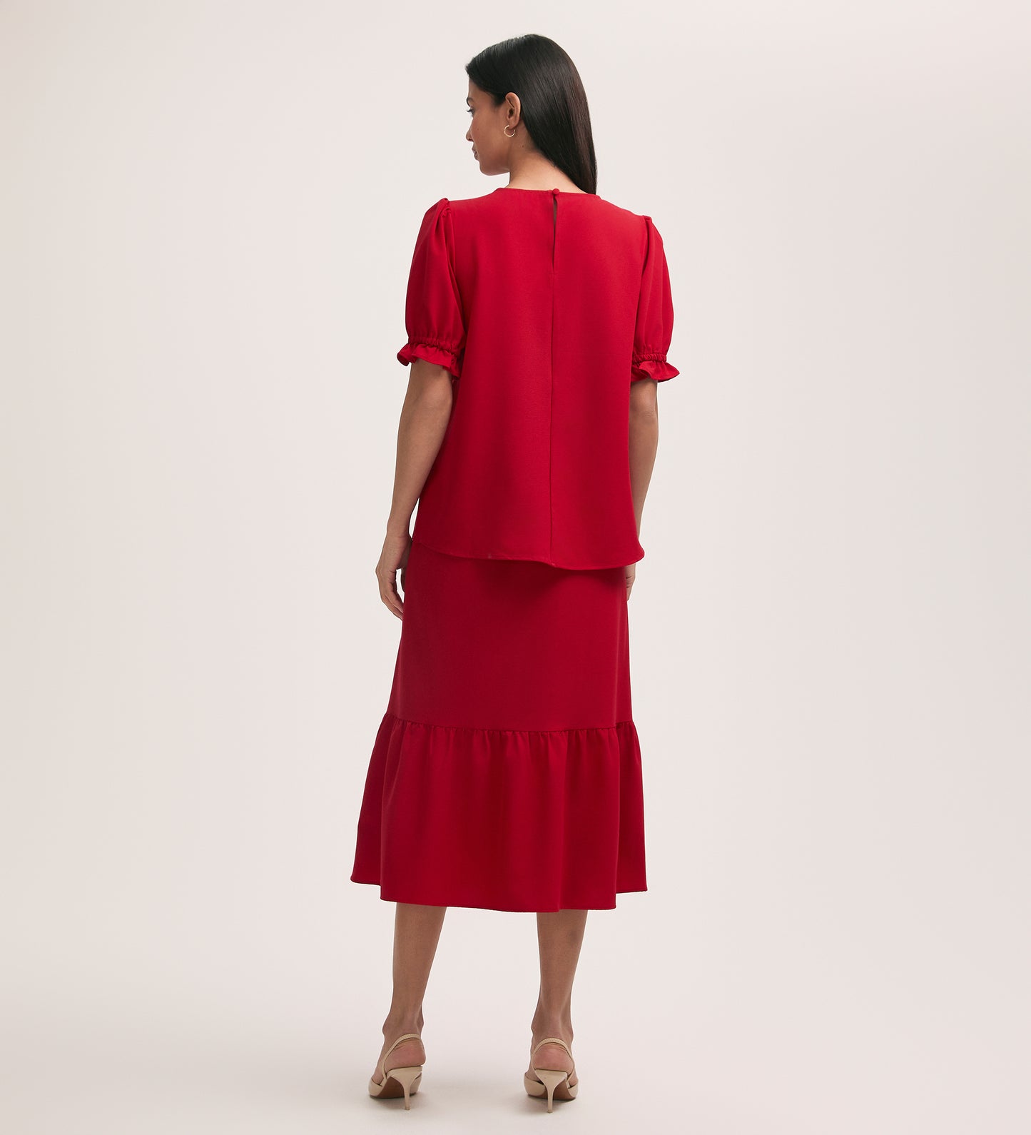 A woman with straight dark hair wears the Charlie Red Crepe T-Shirt with puff sleeves, paired with a matching ruffle-hem midi skirt. She stands facing away against a plain light background.