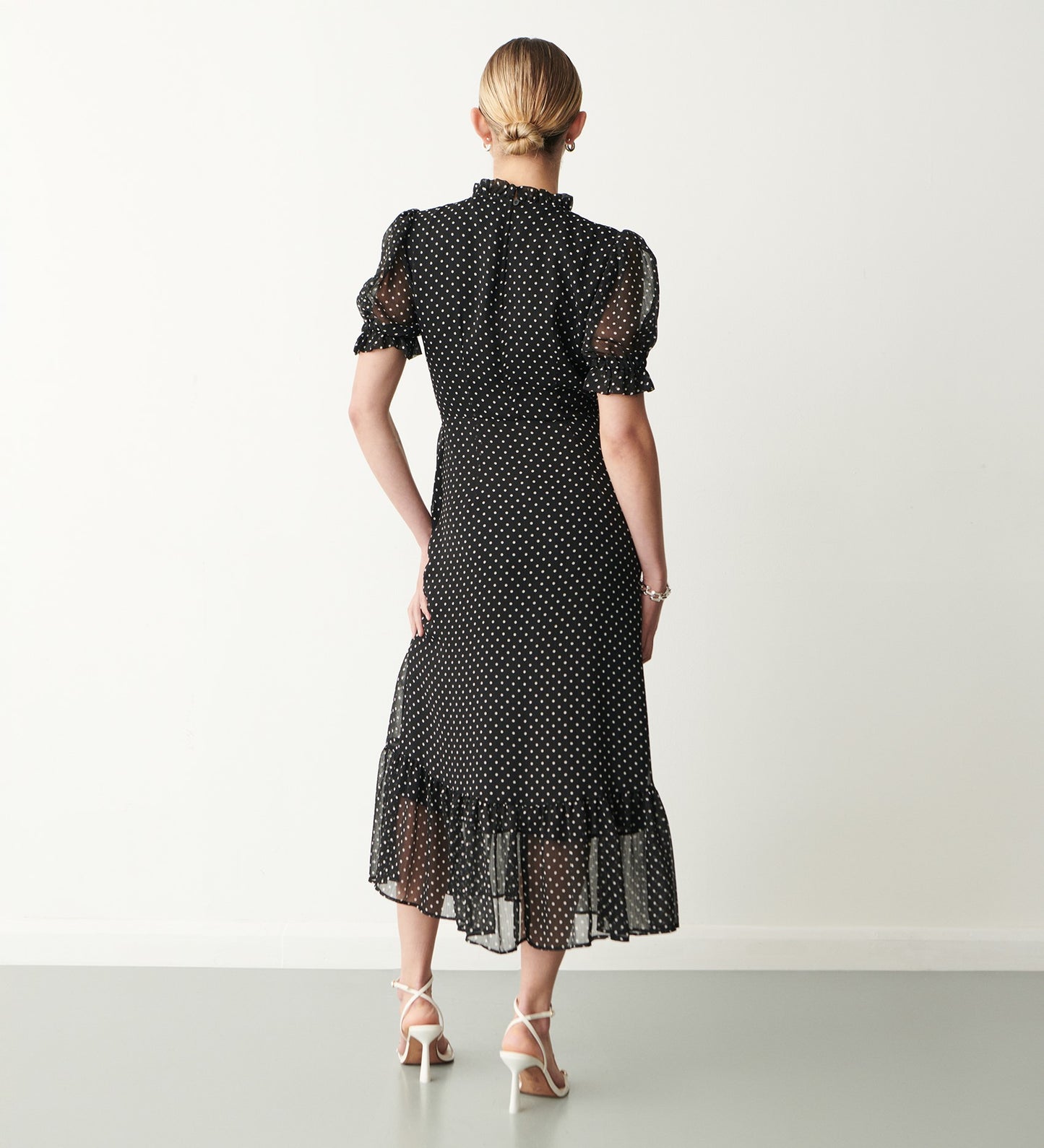 A woman with blonde hair in a low bun models the Camille Chiffon Polka Dot Midi Dress, featuring a ruffled hem and flattering silhouette. She faces away from the camera in white strapped heels against a plain light background.