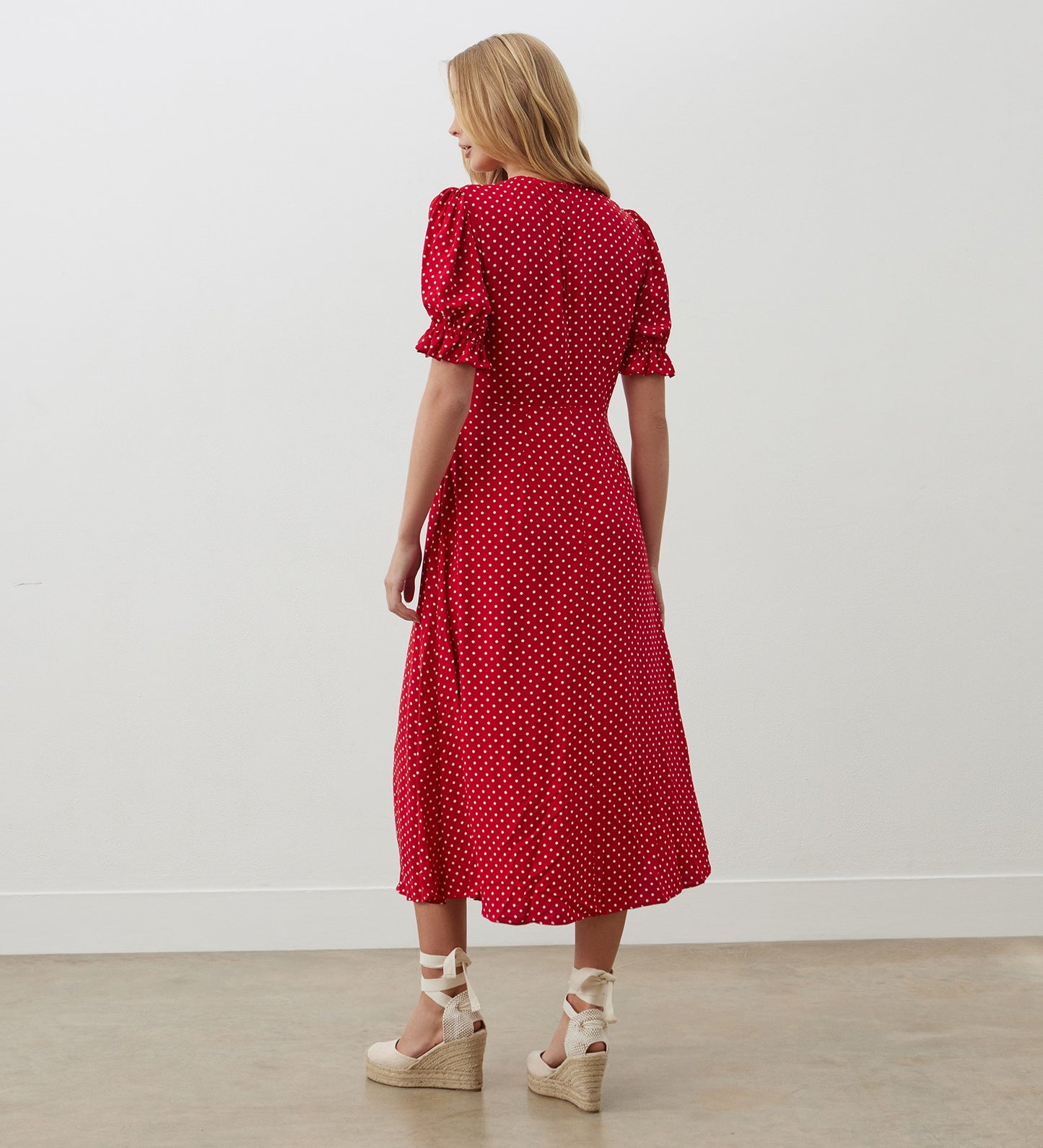 A woman with long blonde hair wears the Mela Red Polka Dot Crepe Midi Dress, featuring an empire waist, and beige wedge sandals, standing with her back to the camera in a minimal indoor setting.