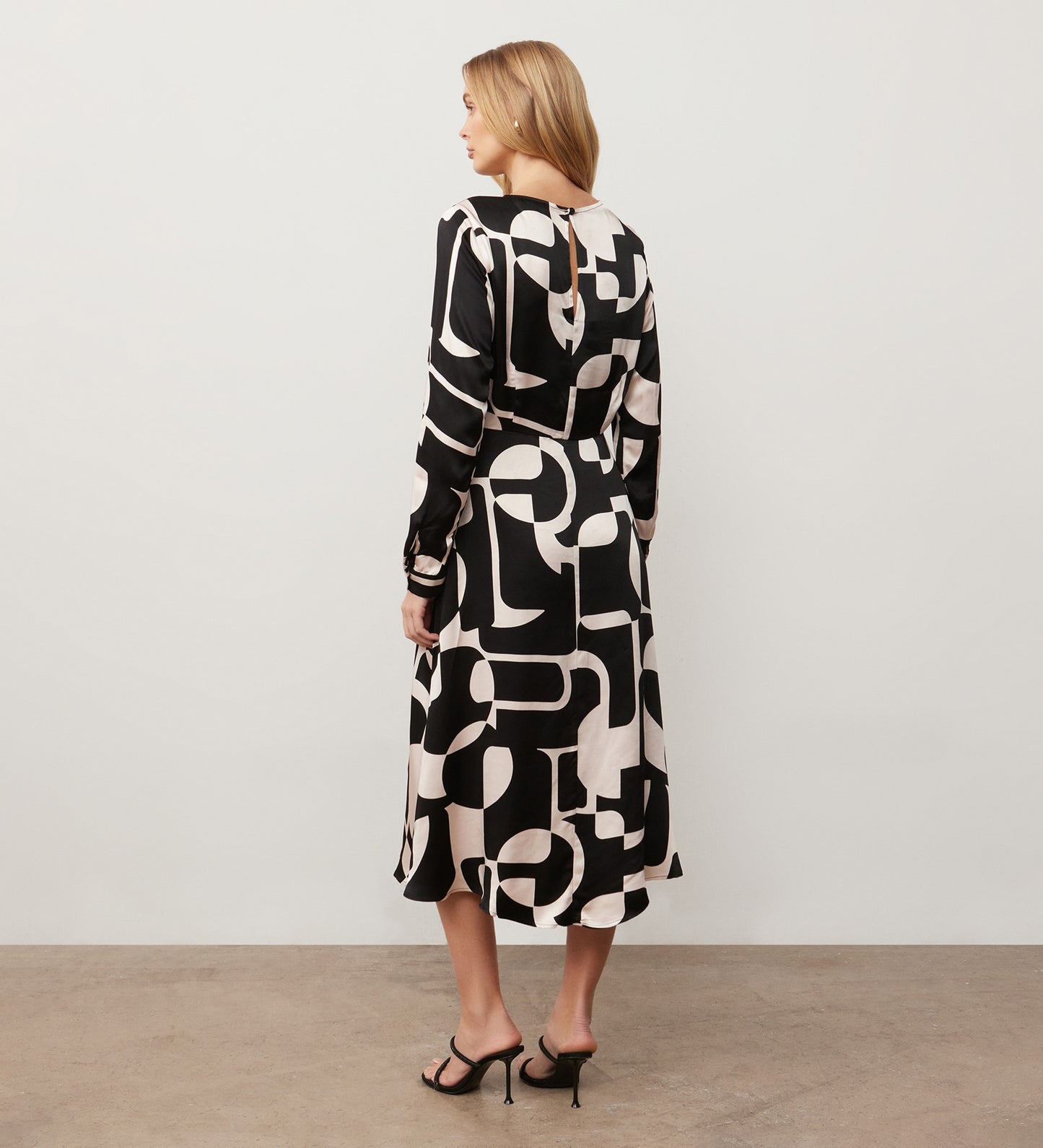 A woman stands facing away, wearing the Annie Black Geo Midi Dress with a tie waist and black heeled sandals, set against a plain light wall and concrete floor.