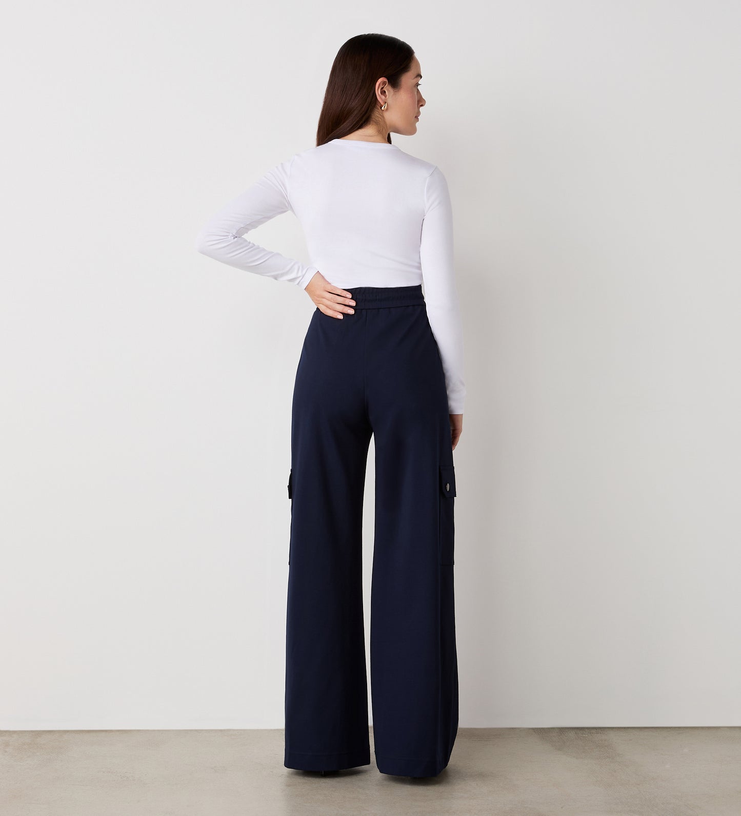 A woman stands with her back to the camera, wearing a fitted white long-sleeve top and Deliah Navy Ponte Jersey Cargo Trousers with a wide-leg, high-waisted fit, set against a plain white wall and concrete floor.