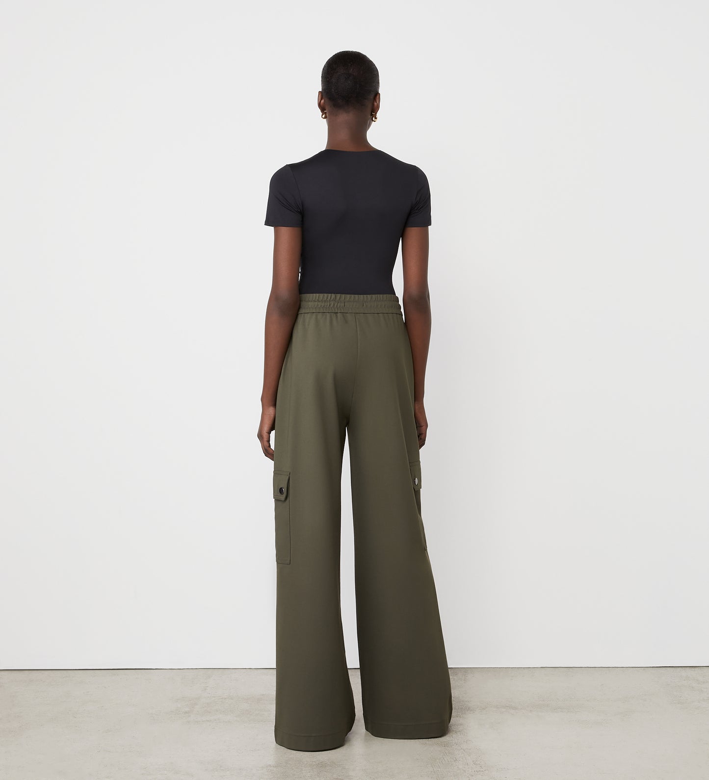 A person faces a white wall, seen from behind, wearing a fitted black short-sleeve top and Deliah Khaki Ponte Jersey Cargo High Waisted Trousers with a wide leg. The minimalist setting includes a plain floor and wall.