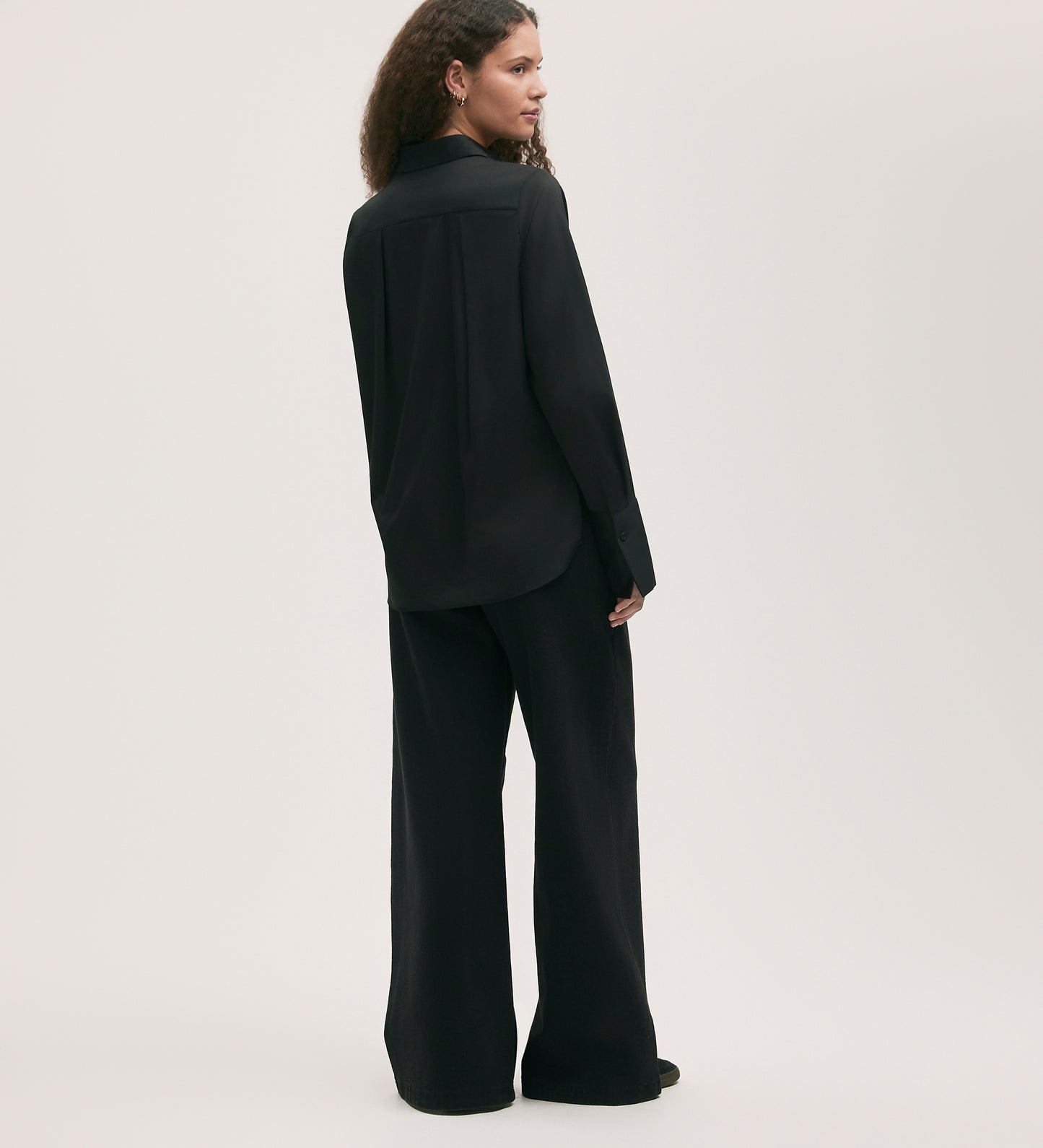 A person with long curly hair wears the Dillon Black Cotton Shirt, featuring a stylish twist, and wide-leg trousers, standing at an angle against a plain light background.