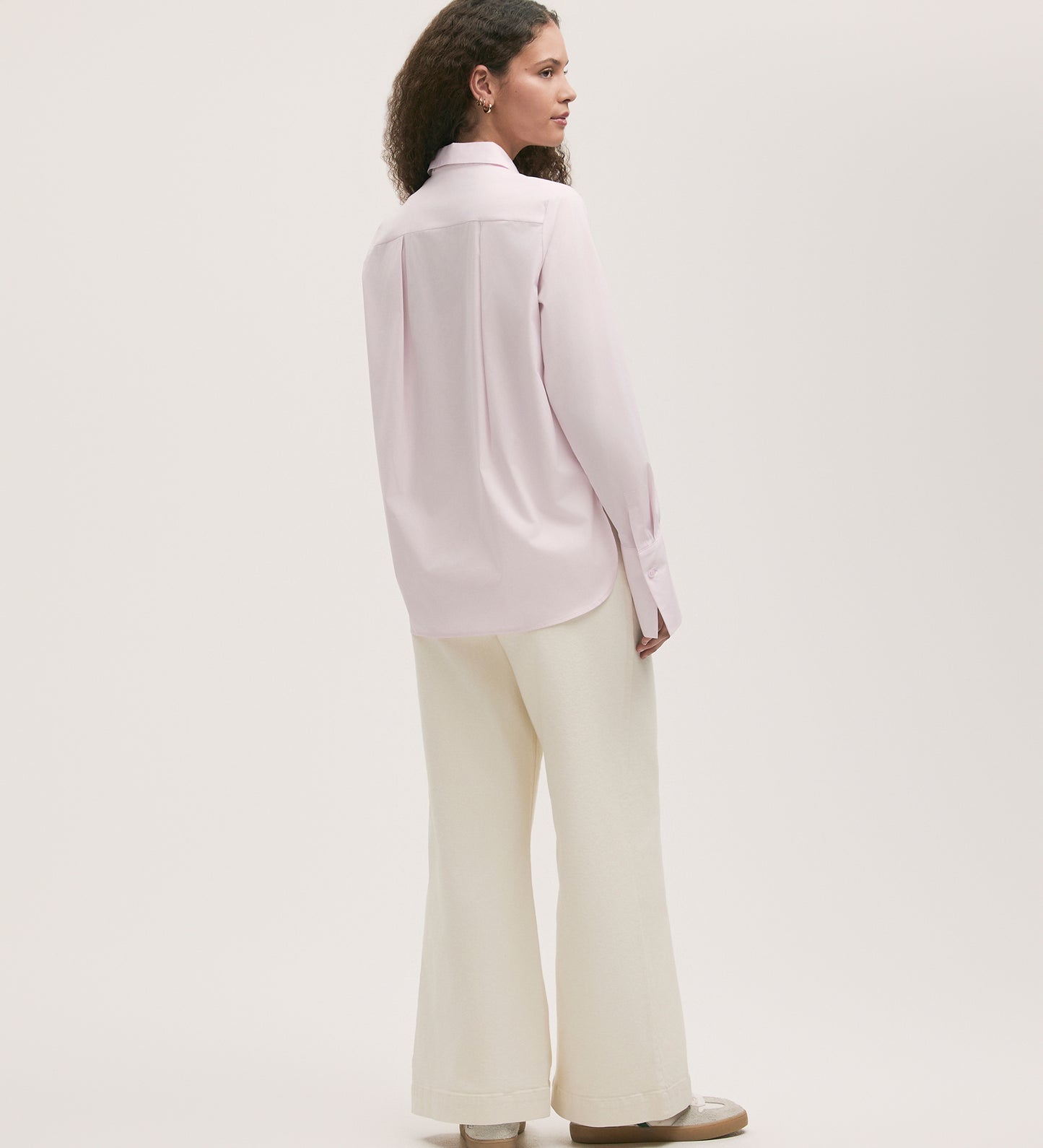A woman with curly brown hair, wearing the Dillon Pink Cotton Shirt (regular fit, long-sleeve) and white wide-leg pants with beige shoes, stands facing away against a plain white background.