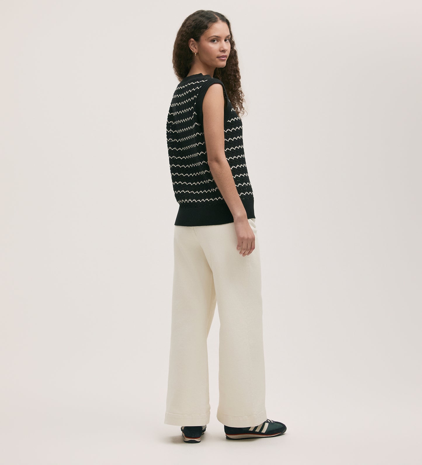 A woman with curly hair wears the Dion Black Crochet Vest, which is sleeveless and features a white zig-zag striped design. She pairs it with wide-leg white pants and black shoes, standing sideways against a plain light background.