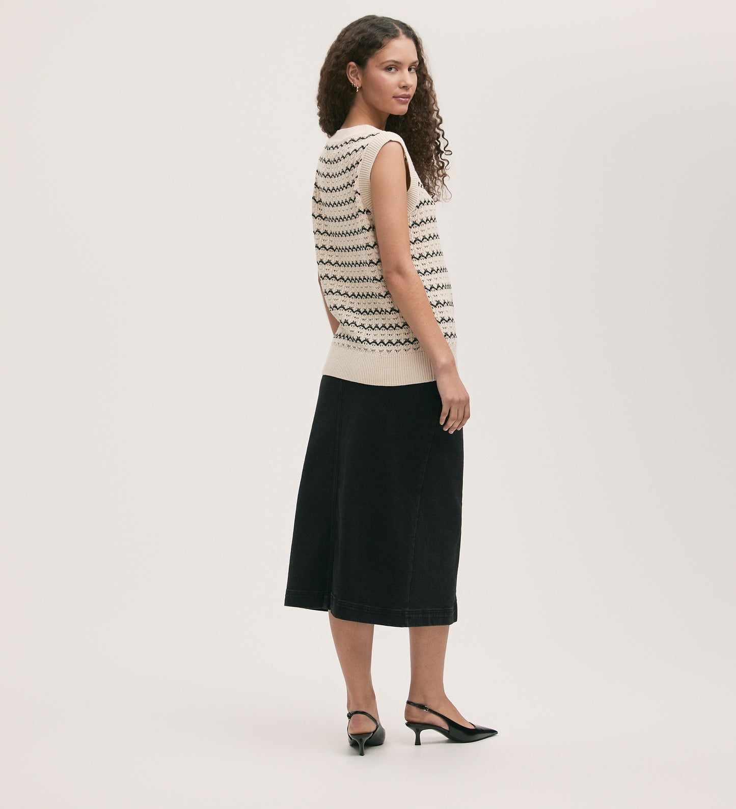 A woman with curly hair wears the Dion Ivory Crochet Vest, styled with a black midi skirt and heeled shoes, standing sideways and looking over her shoulder against a plain light background.