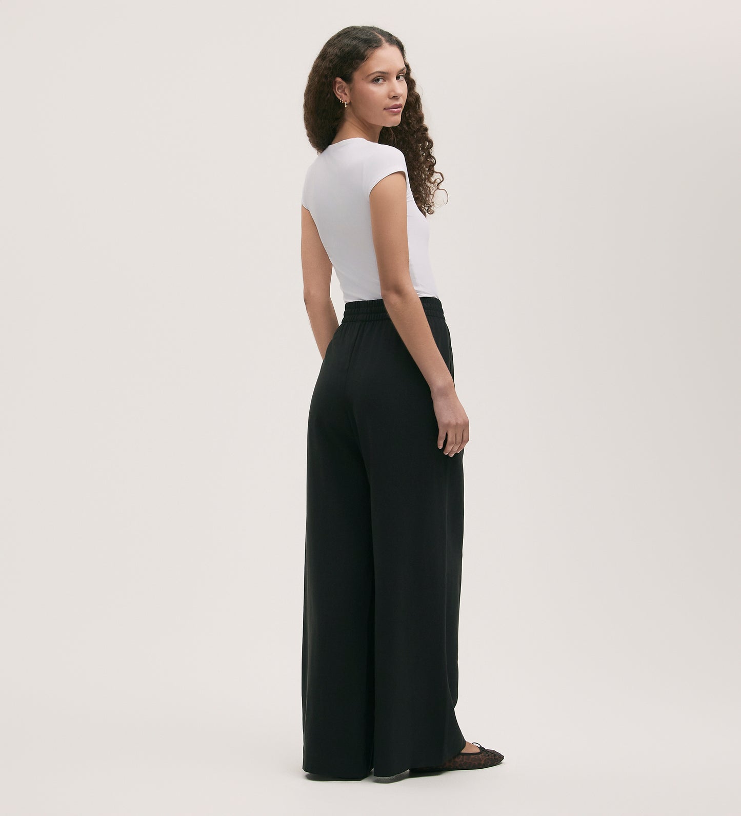 A woman with long curly hair looks over her shoulder, wearing a fitted white short-sleeve top and the Dixie Black Wide Leg Trousers, which feature a high waist and elasticated waistband, paired with black shoes against a light background.